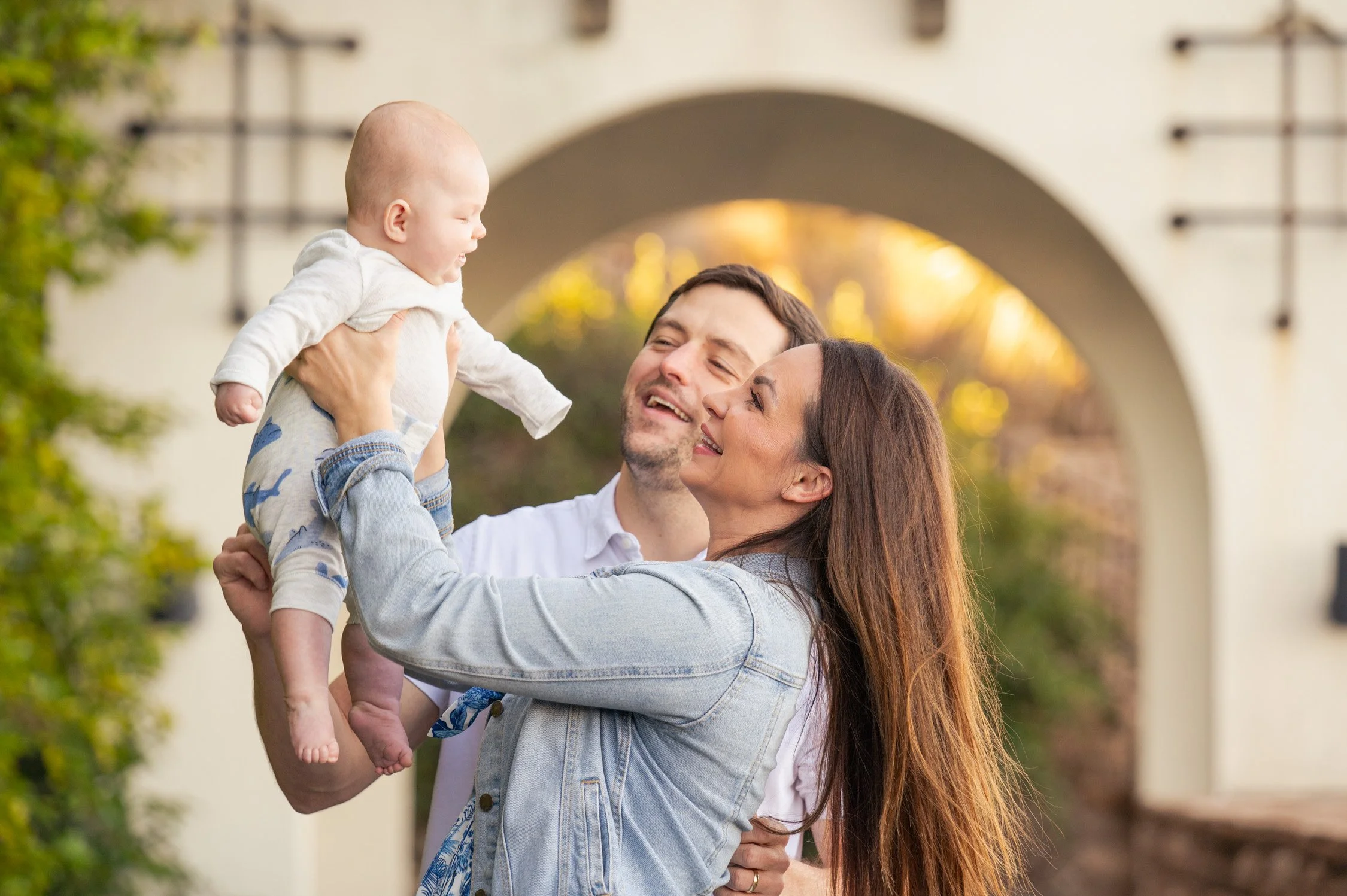 A family of three, a mother, father, and a baby, smiling and playing outdoors during sunset, with the mother lifting the baby and the father standing close by.