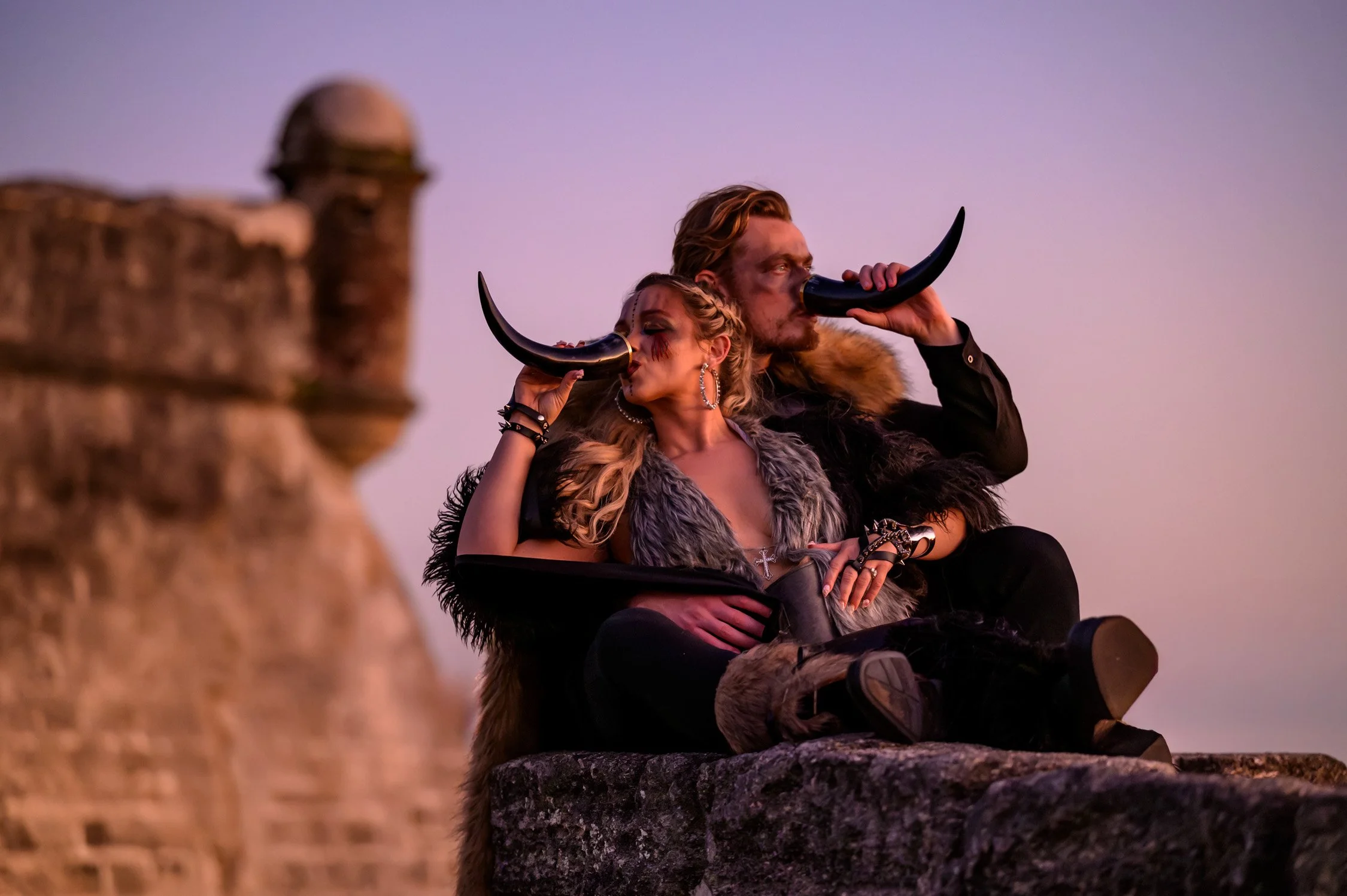 Two people, a woman and a man, sitting on a stone surface, both holding black horns to their mouths. They are dressed in dark, fur-lined clothing with accessories, and appear to be looking into the distance during sunset with a fortification wall in 