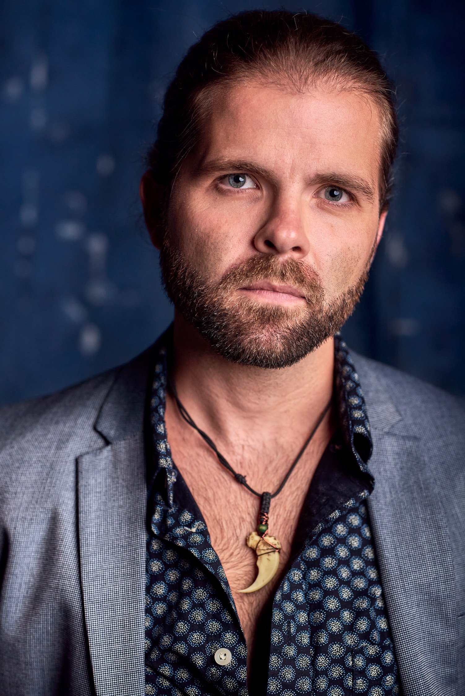 A man with long hair and a beard wearing a gray blazer, black patterned shirt, and a necklace with a fang-shaped pendant.