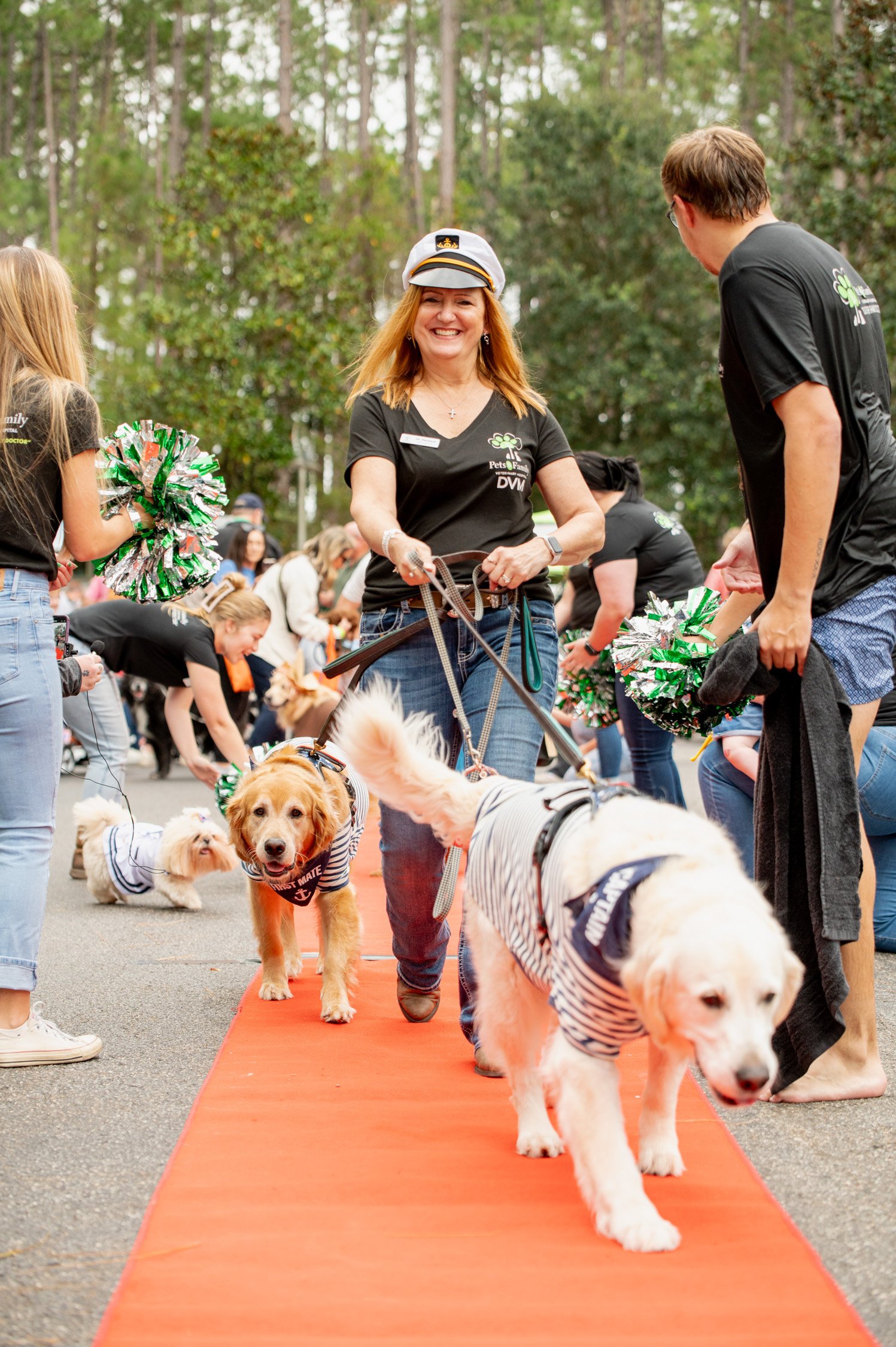 People and dogs participating in a pet parade on a red carpet outdoors, with green trees in the background. A woman in a captain's hat smiles with dogs dressed in striped shirts.