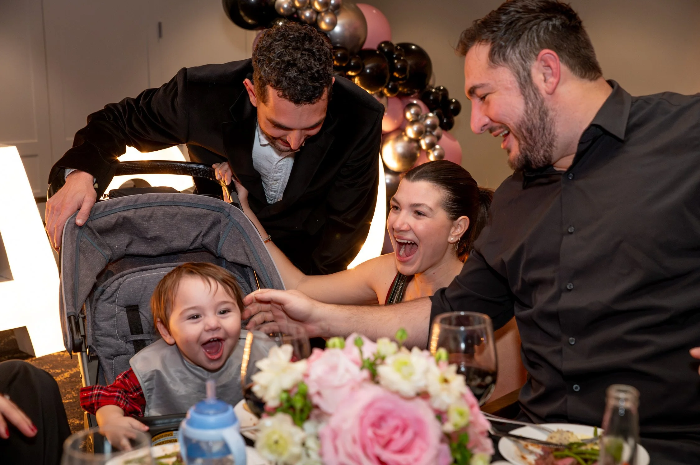A group of four people, including two men, a woman, and a young child in a stroller, are gathered around a table celebrating. They are smiling and appear to be happy, with the woman and one of the men reaching out to the child. There are flowers and 
