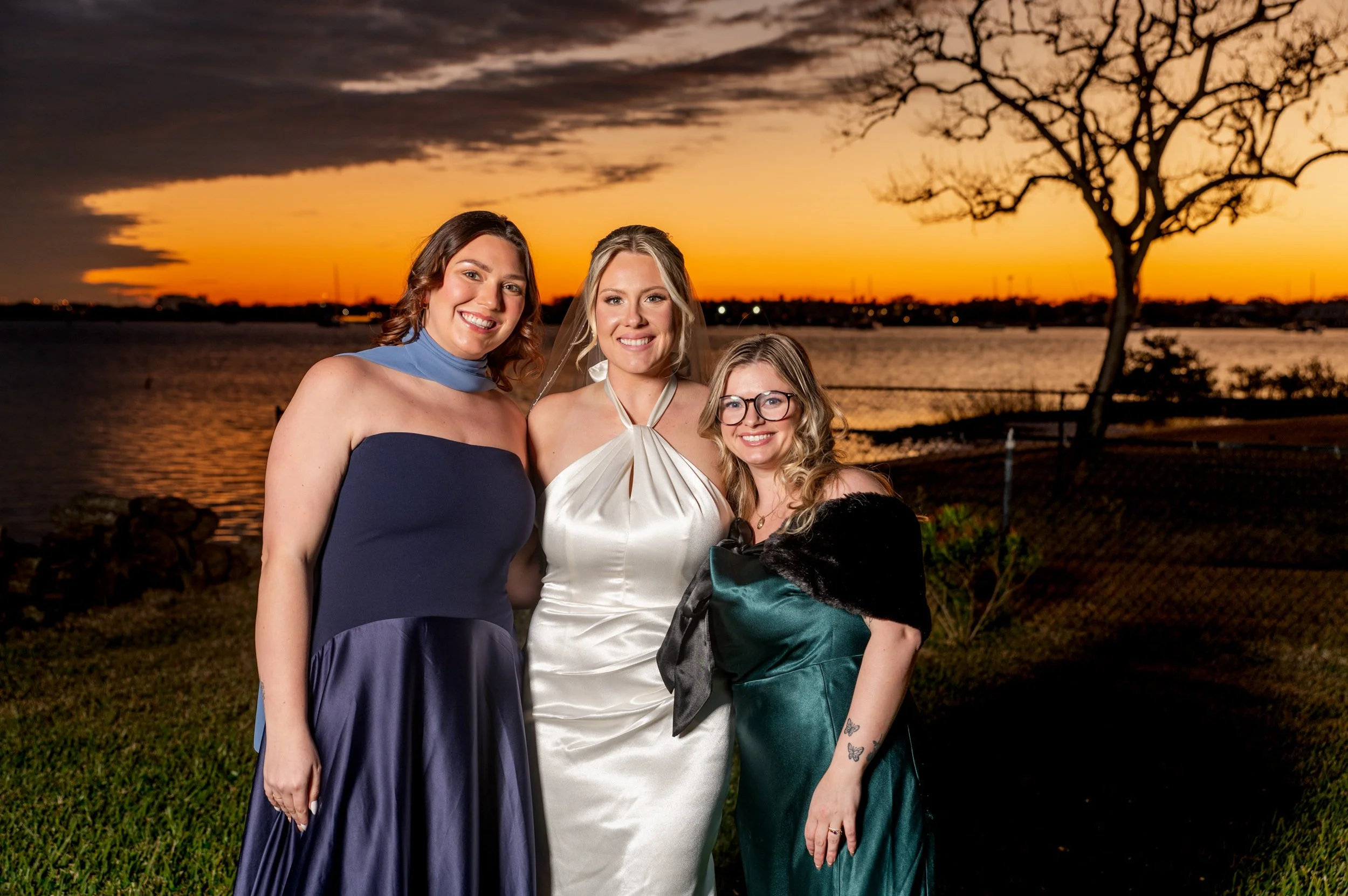 Three women in formal dresses smiling outdoors during sunset, with a river, a tree, and a fence in the background.