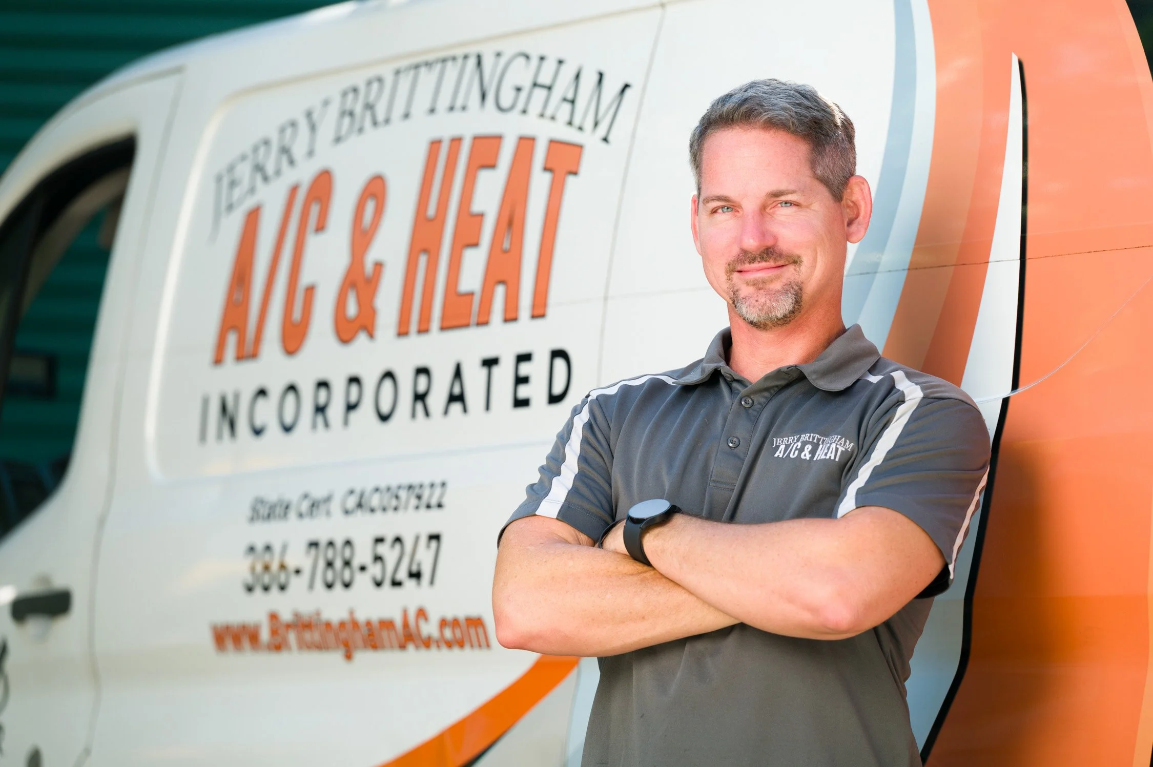 A man with crossed arms standing in front of a branded company van, wearing a gray polo shirt with the company logo, smiling slightly.