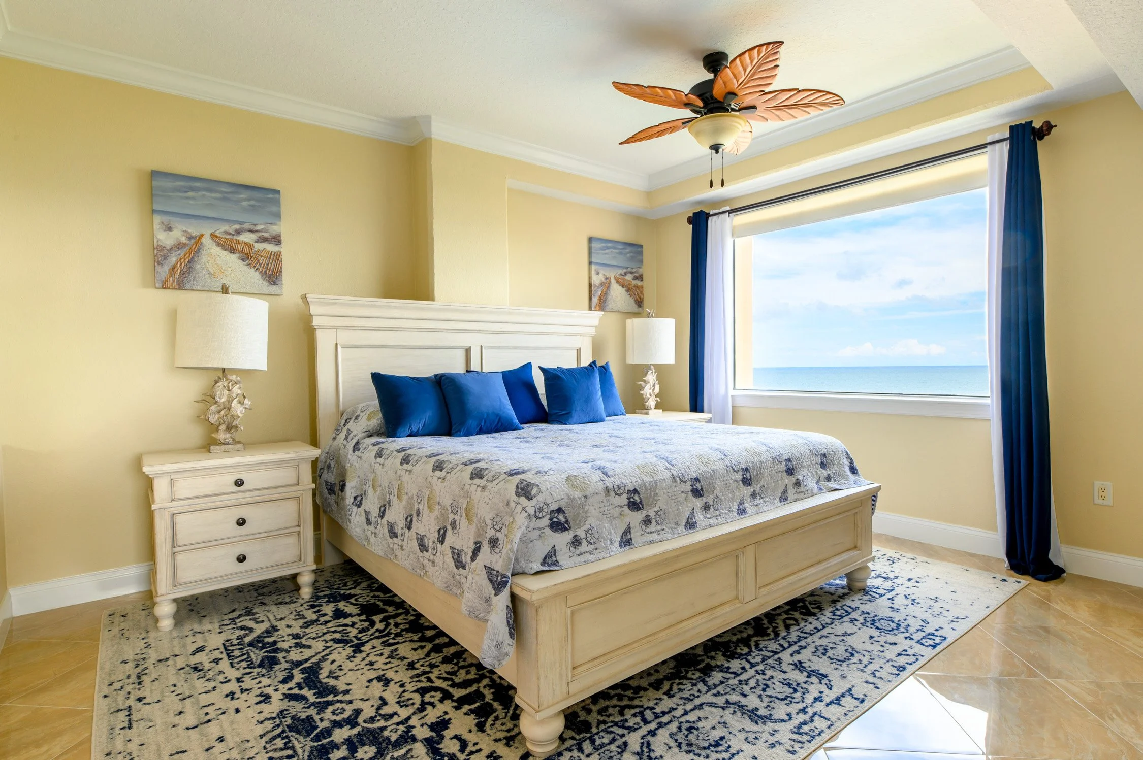 A bedroom with a large window showing the ocean view, a white bed with blue pillows, two nightstands with lamps, a ceiling fan with leaf-shaped blades, and beach-themed artwork on pale yellow walls.