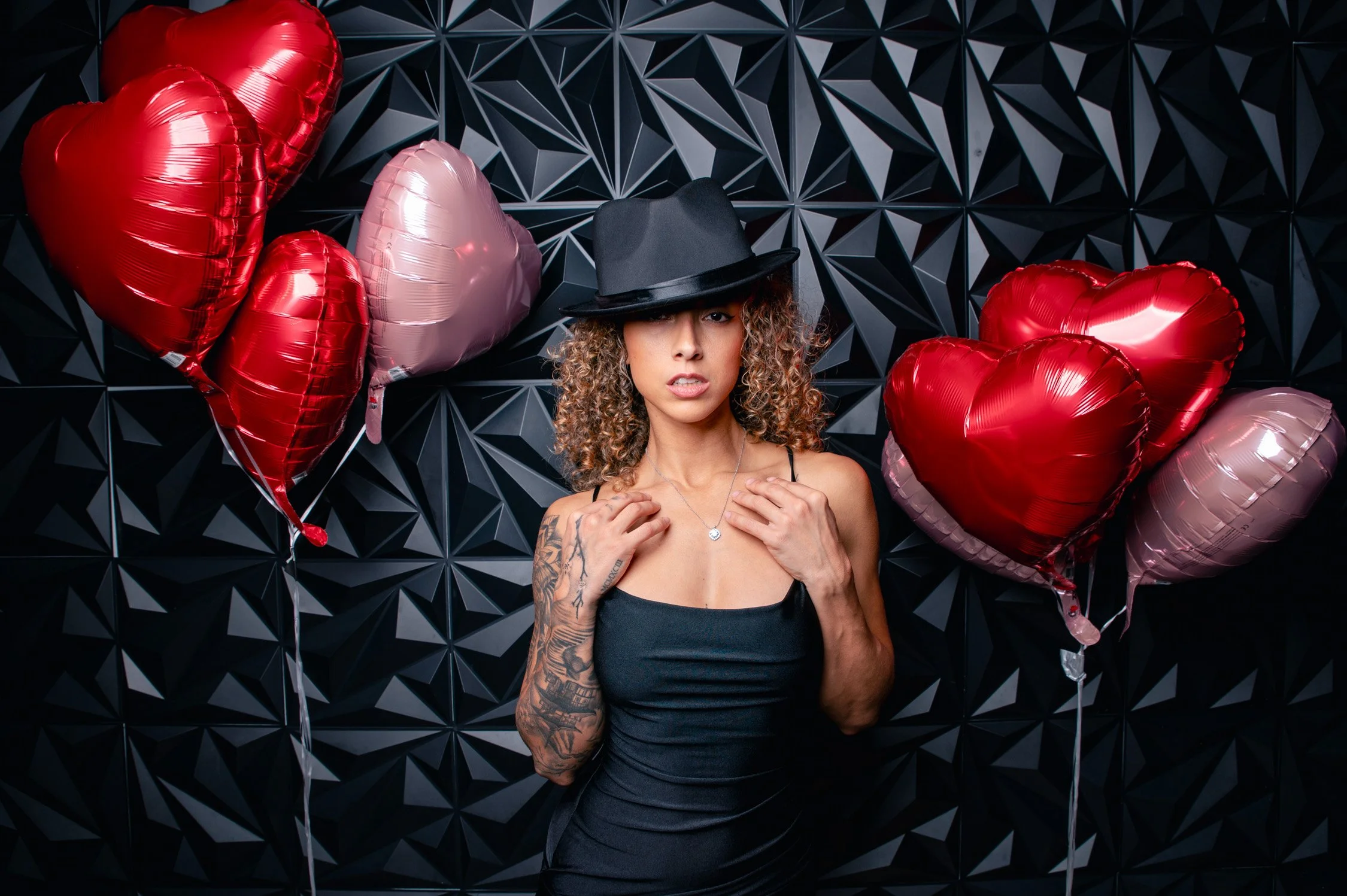 A woman with curly hair wearing a black dress and a black hat, standing in front of a black geometric patterned wall, surrounded by red and pink heart-shaped balloons.