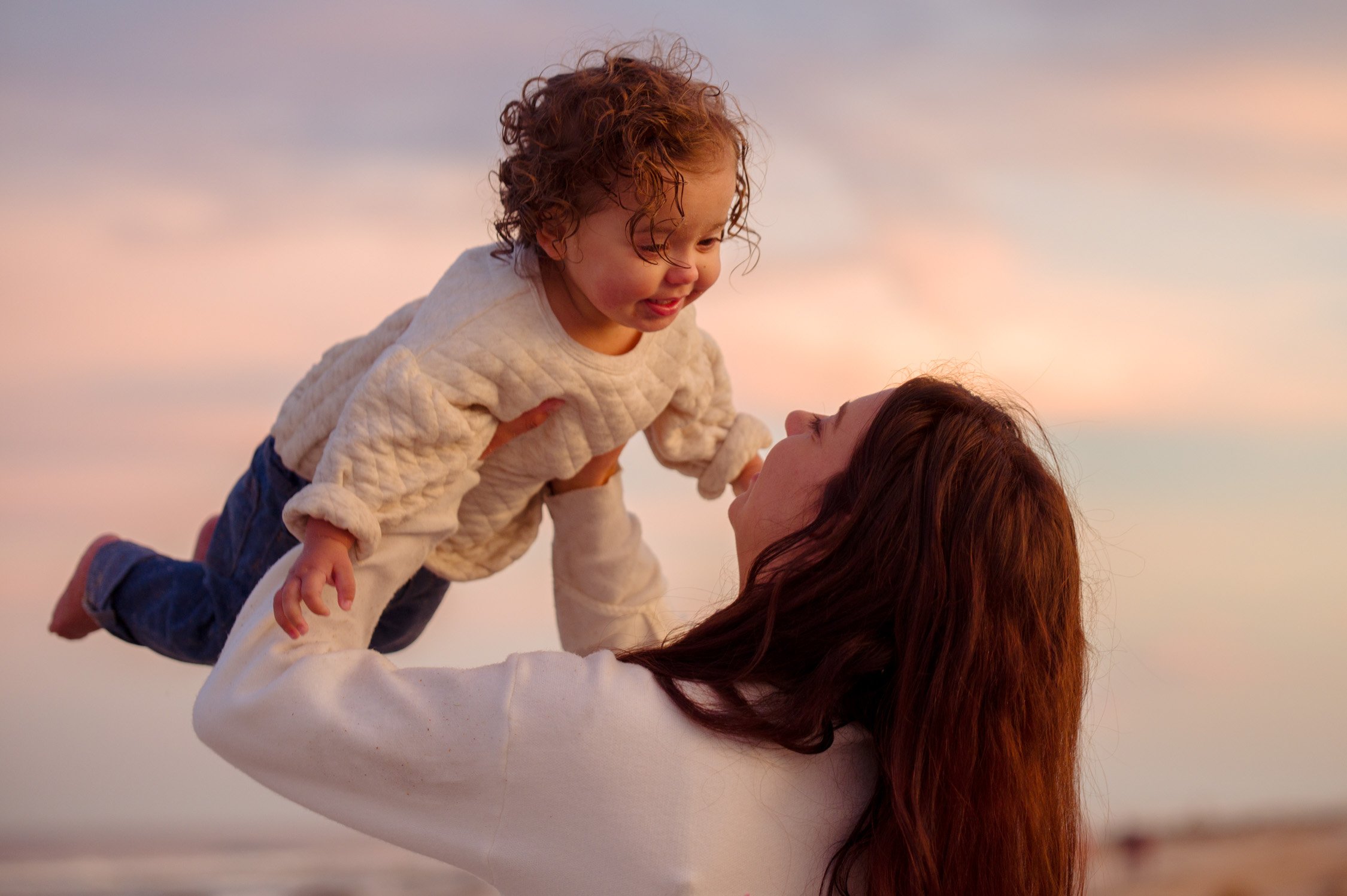 A woman with red hair lifting a curly-haired child with light brown hair, both smiling and looking at each other on a beach during sunset.