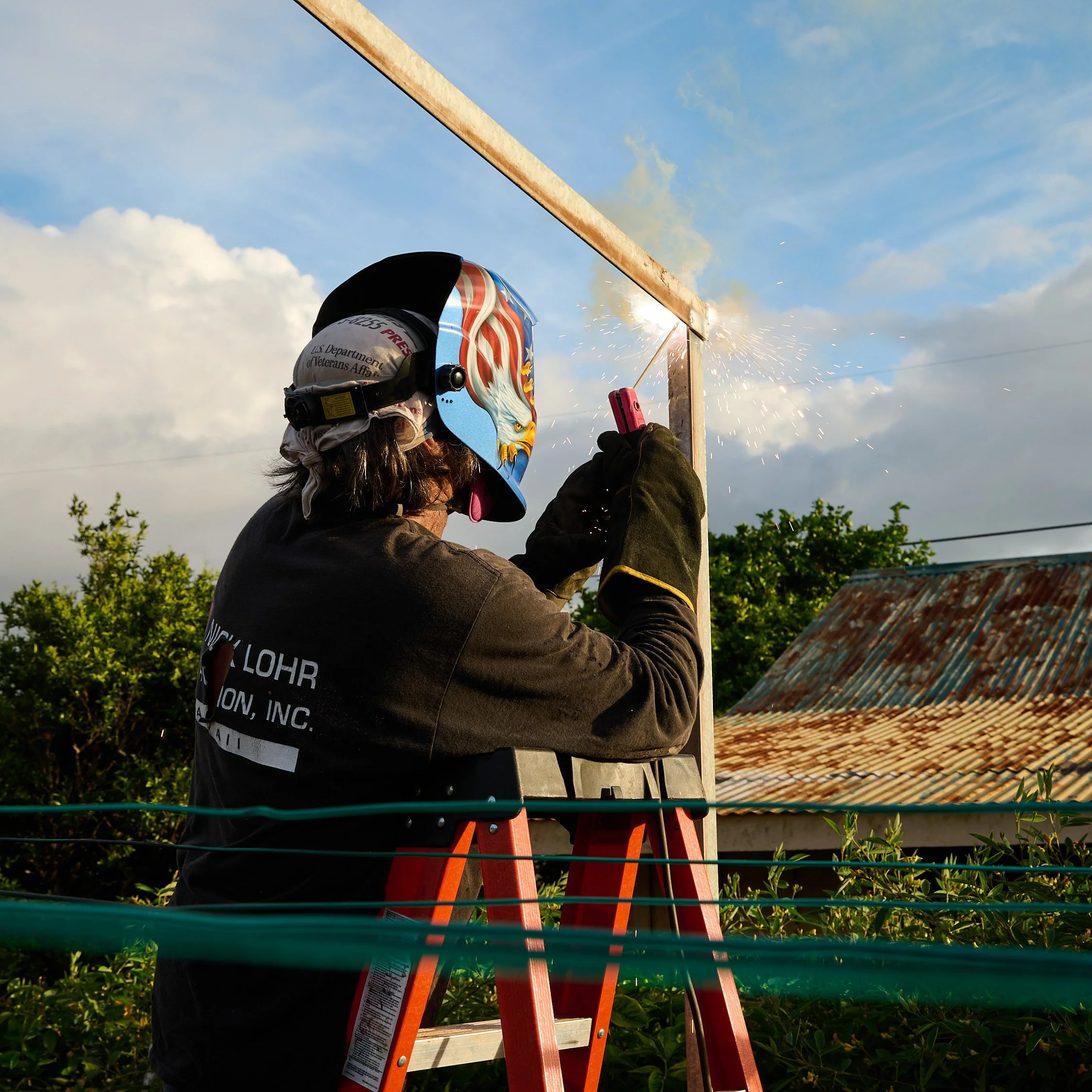 A person welding outdoors on a ladder, wearing a helmet with patriotic graphics, welding gloves, and dark clothing, welding a metal frame during daylight.
