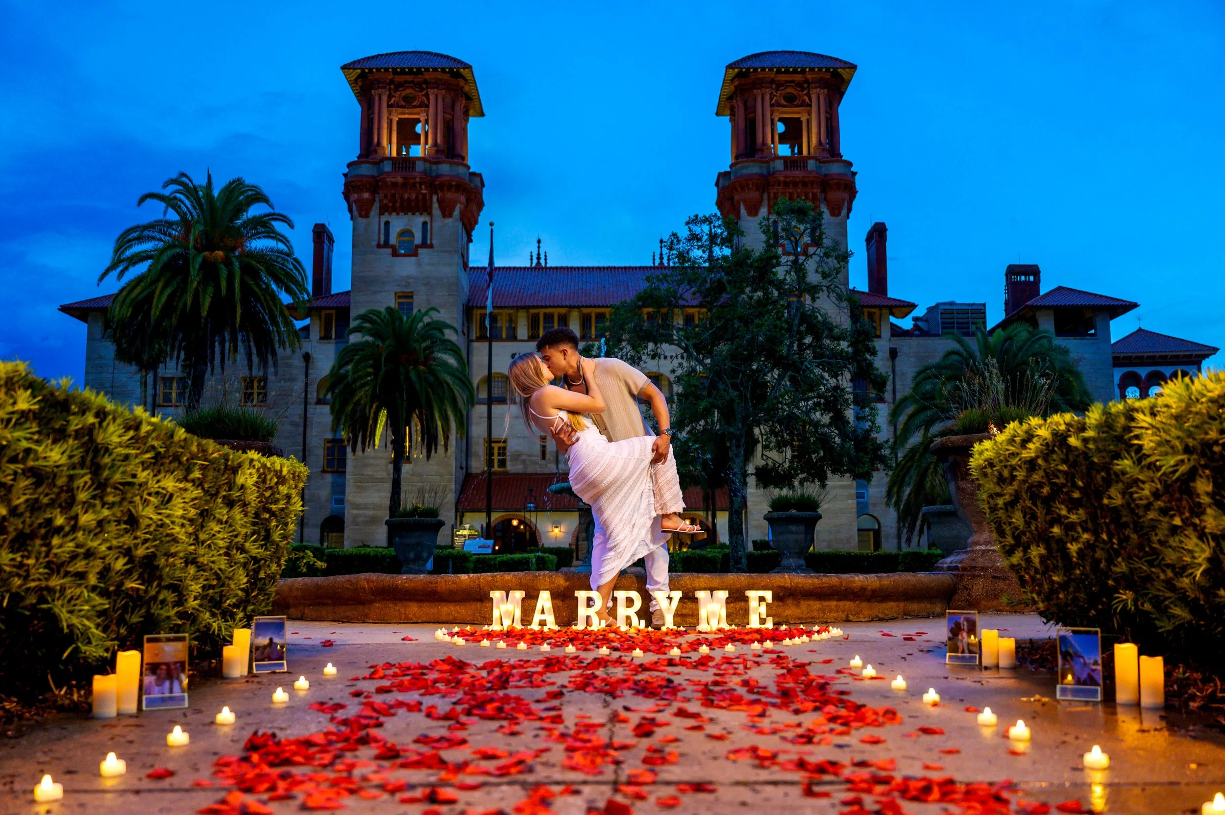 A couple sharing a romantic kiss, with the man lifting the woman in front of a large castle-like building at dusk, surrounded by palm trees, candles, and rose petals on the ground, with illuminated letters spelling 'MARRY ME' in front of them.