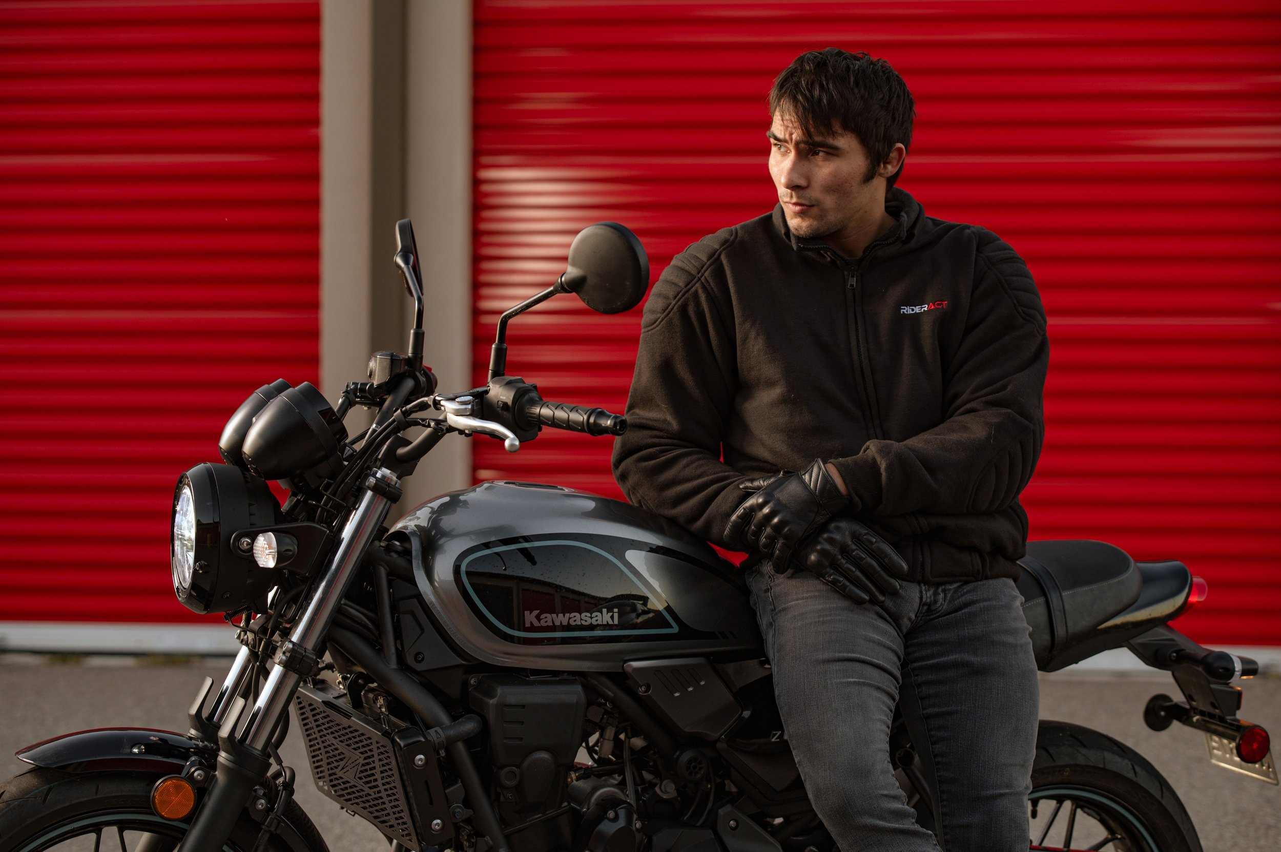 A man wearing a black jacket and gloves sitting on a black Kawasaki motorcycle in front of a red industrial door.