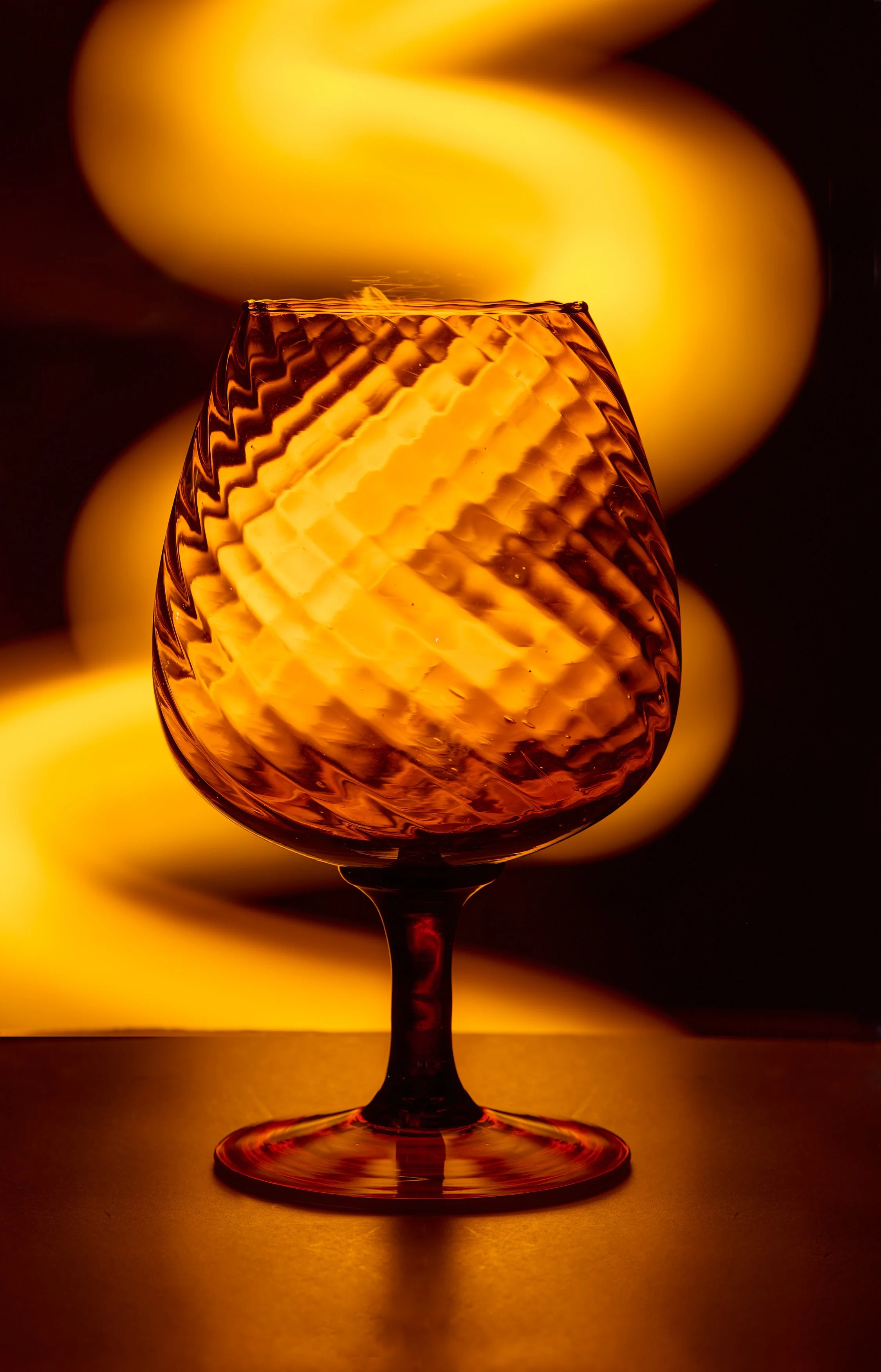A close-up photo of an amber-colored glass with a textured, spiral pattern, lit from behind, with blurred yellow-orange shapes in the background.