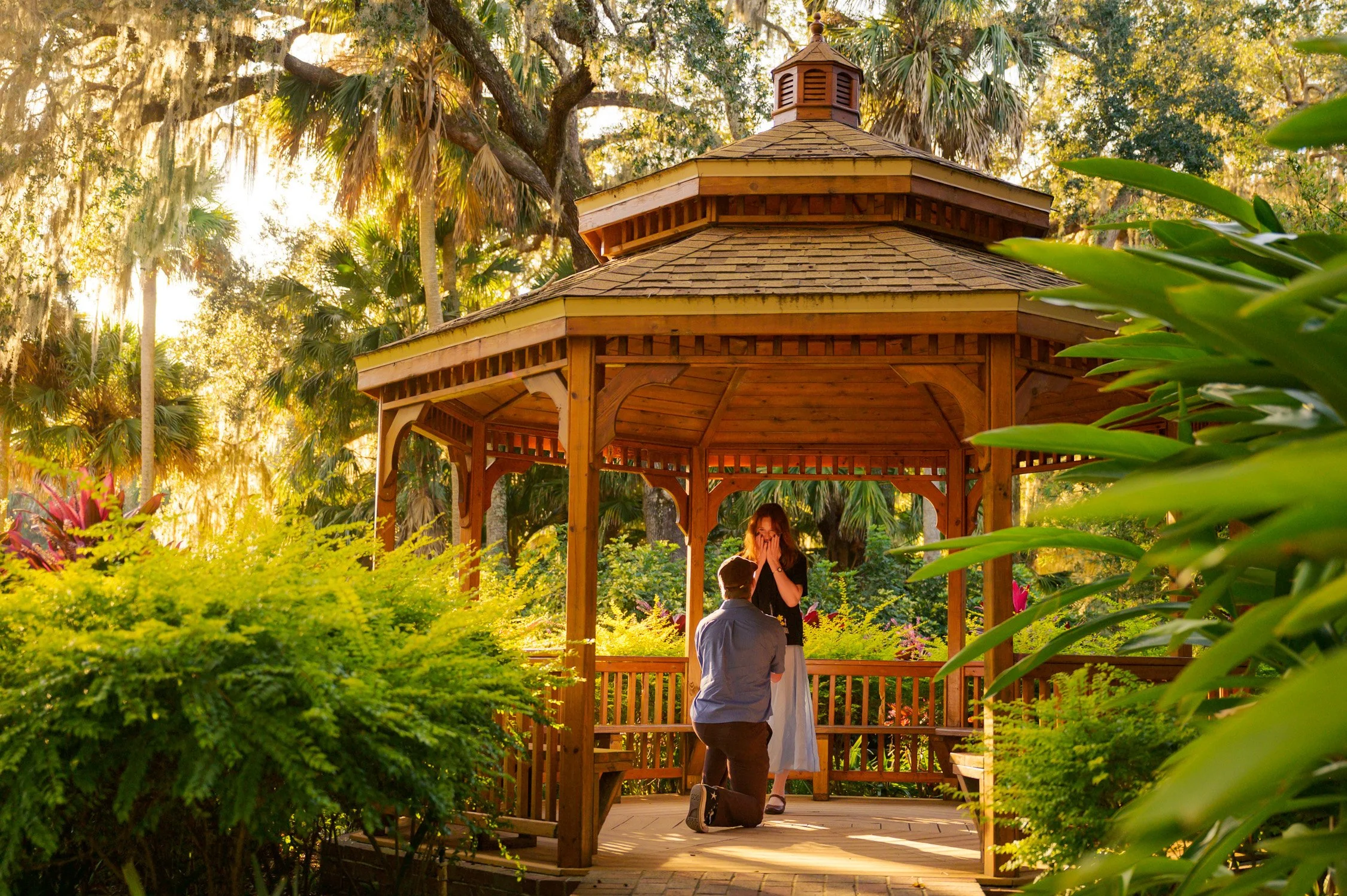 A couple is in a park, with the man kneeling before the woman who covers her face, inside a wooden gazebo surrounded by lush greenery and trees during sunset.