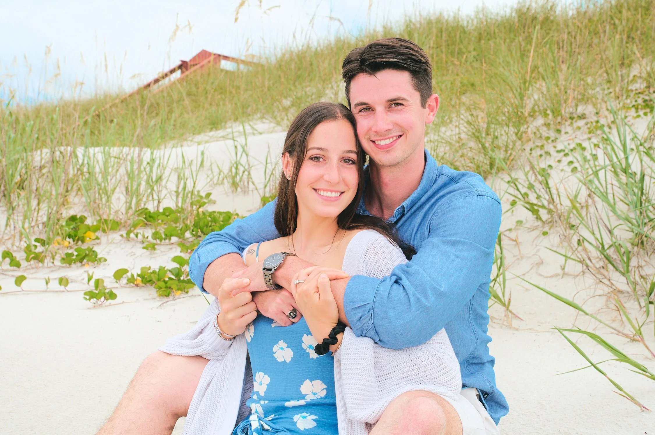 A young couple sitting on a white sandy beach, smiling at the camera, with green grasses and a wooden boardwalk in the background.
