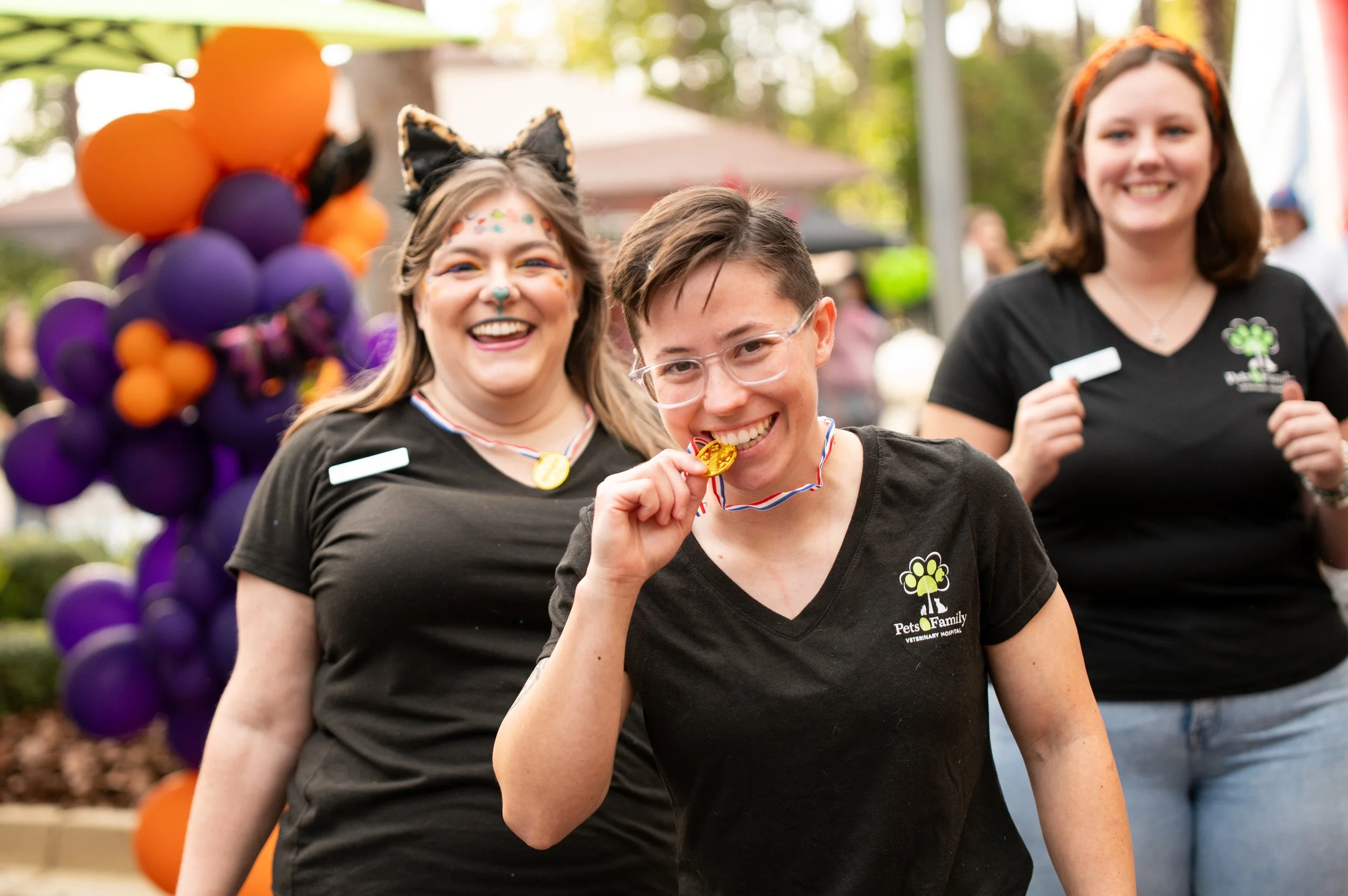 Three women celebrating outdoors, with the woman in the front biting a gold medal, wearing glasses and a black T-shirt from Pets & Family Veterinarian Hospital. The woman to her left has face paint, cat ears headband, and festive makeup. The woman to the right holds a white card and has a big smile. Decorations include purple and orange balloons.