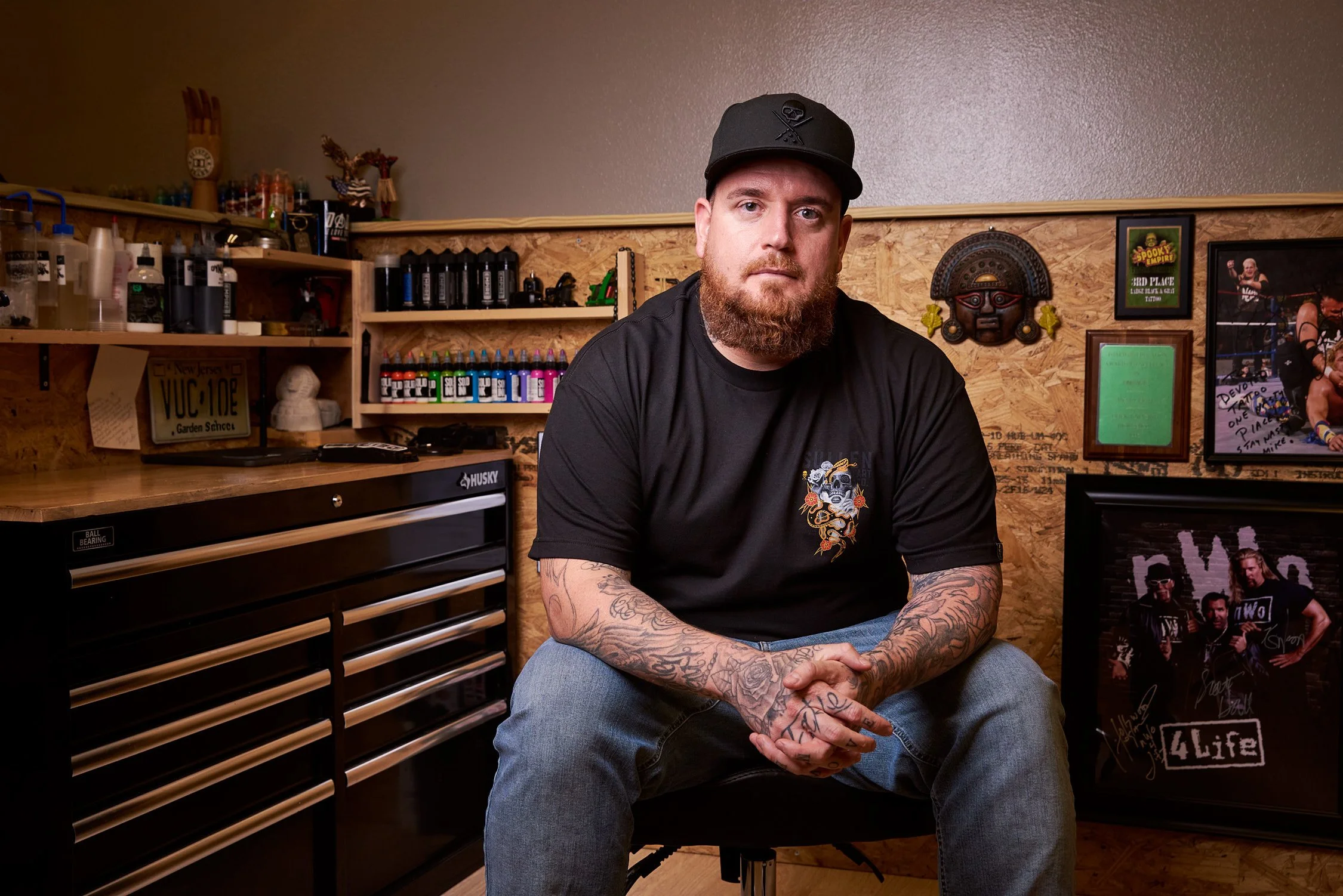 A man with a beard, tattoos, and wearing a black cap and black t-shirt sitting in a workshop with shelves of paint bottles, a Husky tool chest, and framed pictures and art on the wood-paneled wall behind him.
