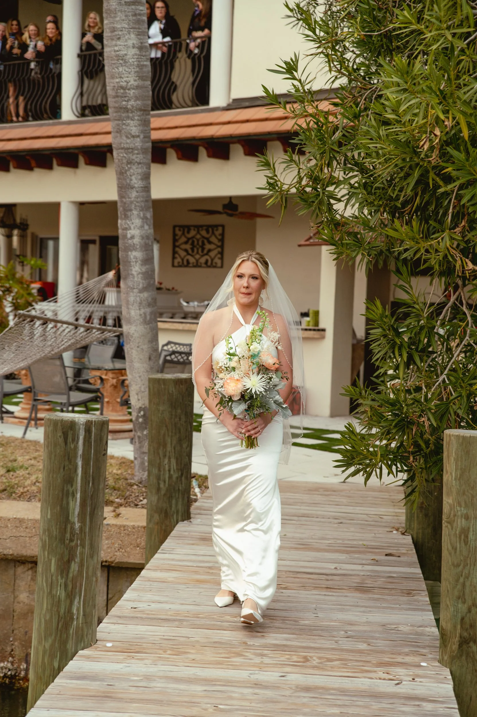 Bride walking on a wooden dock, holding a bouquet of flowers, wearing a satin wedding dress and veil, with a mansion and a group of guests watching from the balcony in the background.
