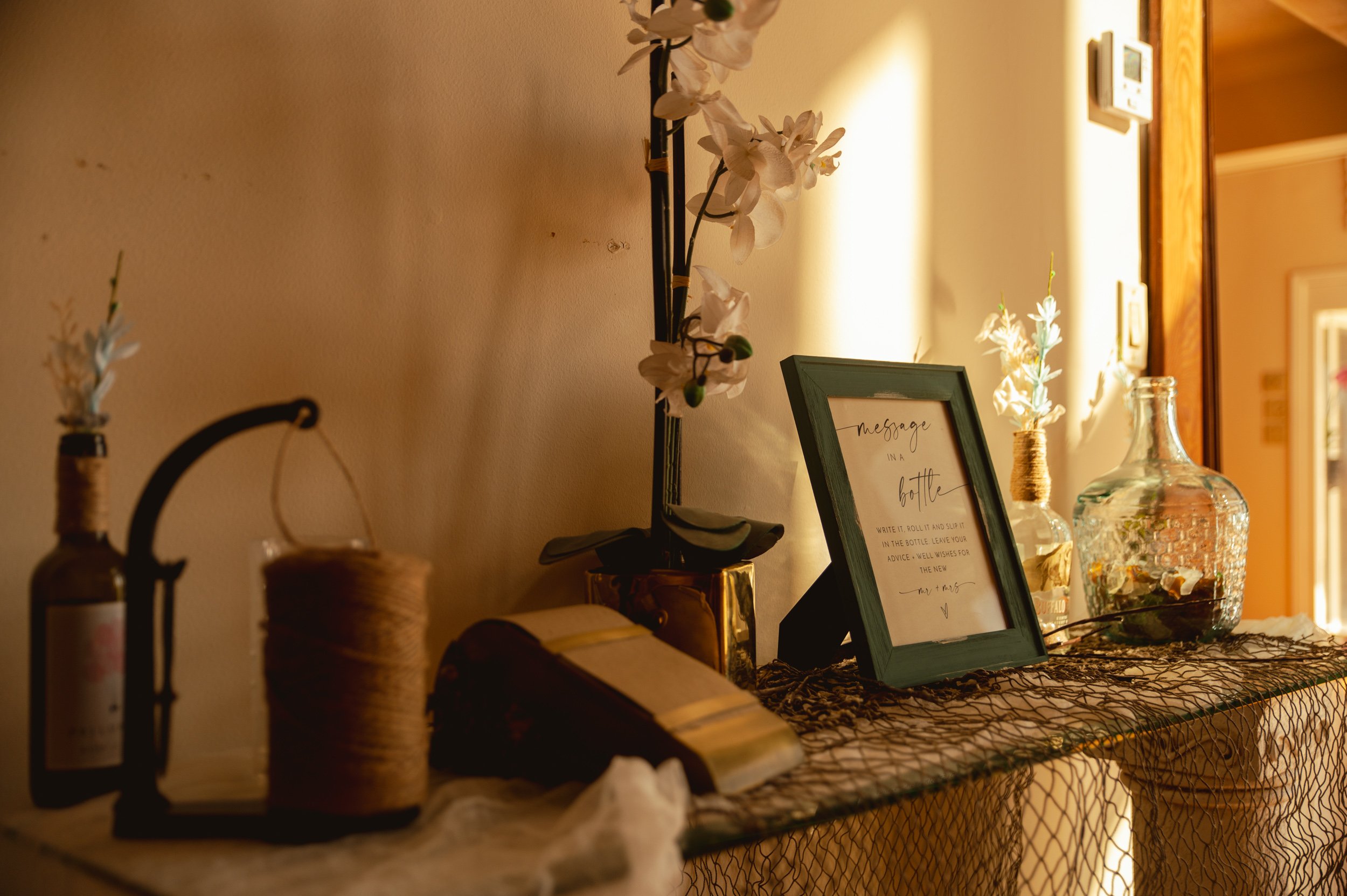 Decorative table with flowers in bottles, a framed message, a book, and a glass jar, illuminated by warm lighting.