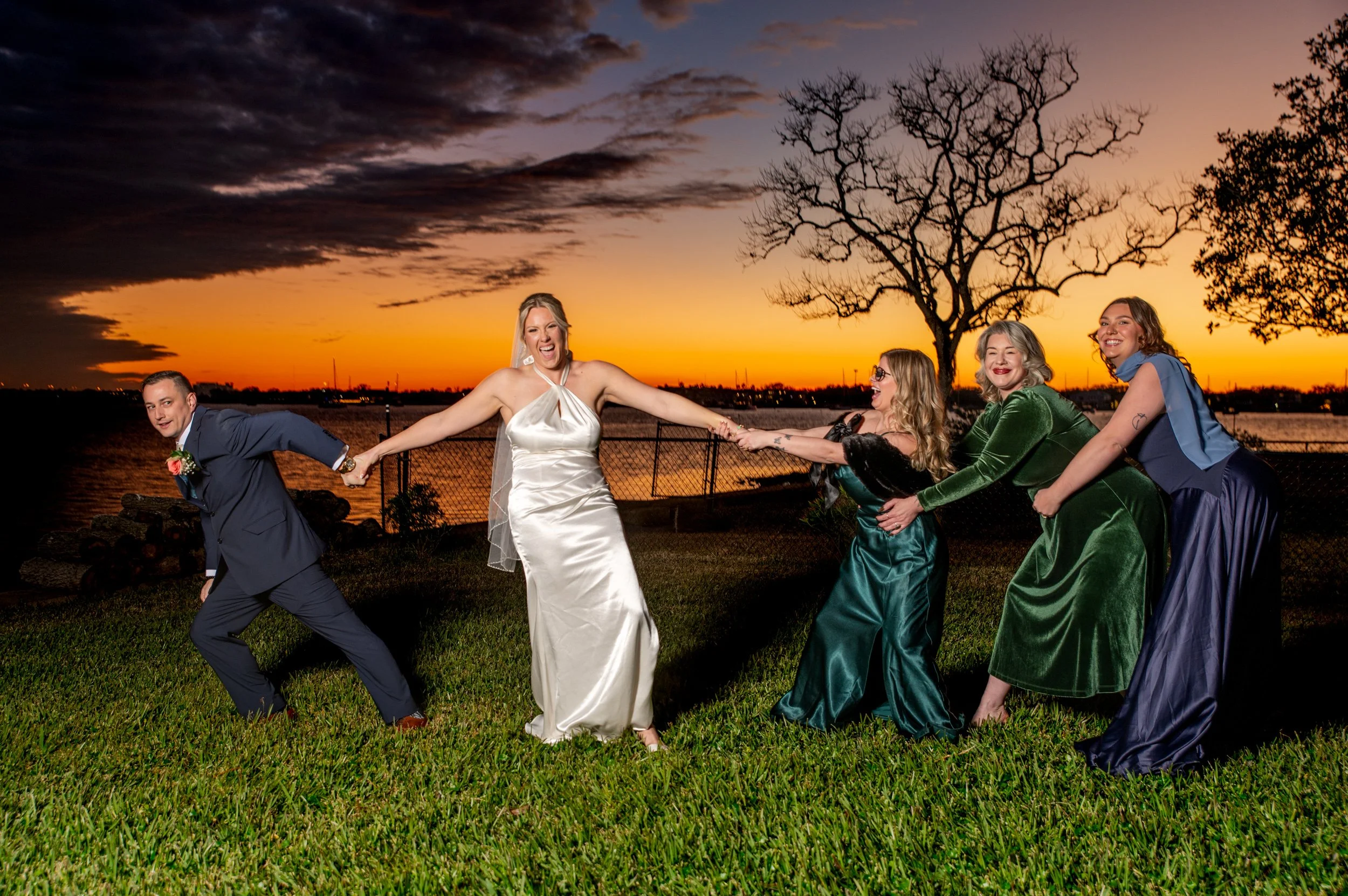 A group of five people, including a bride in a white dress and four women in colorful dresses, holding hands and pulling each other on a grassy area at sunset with a river and trees in the background.