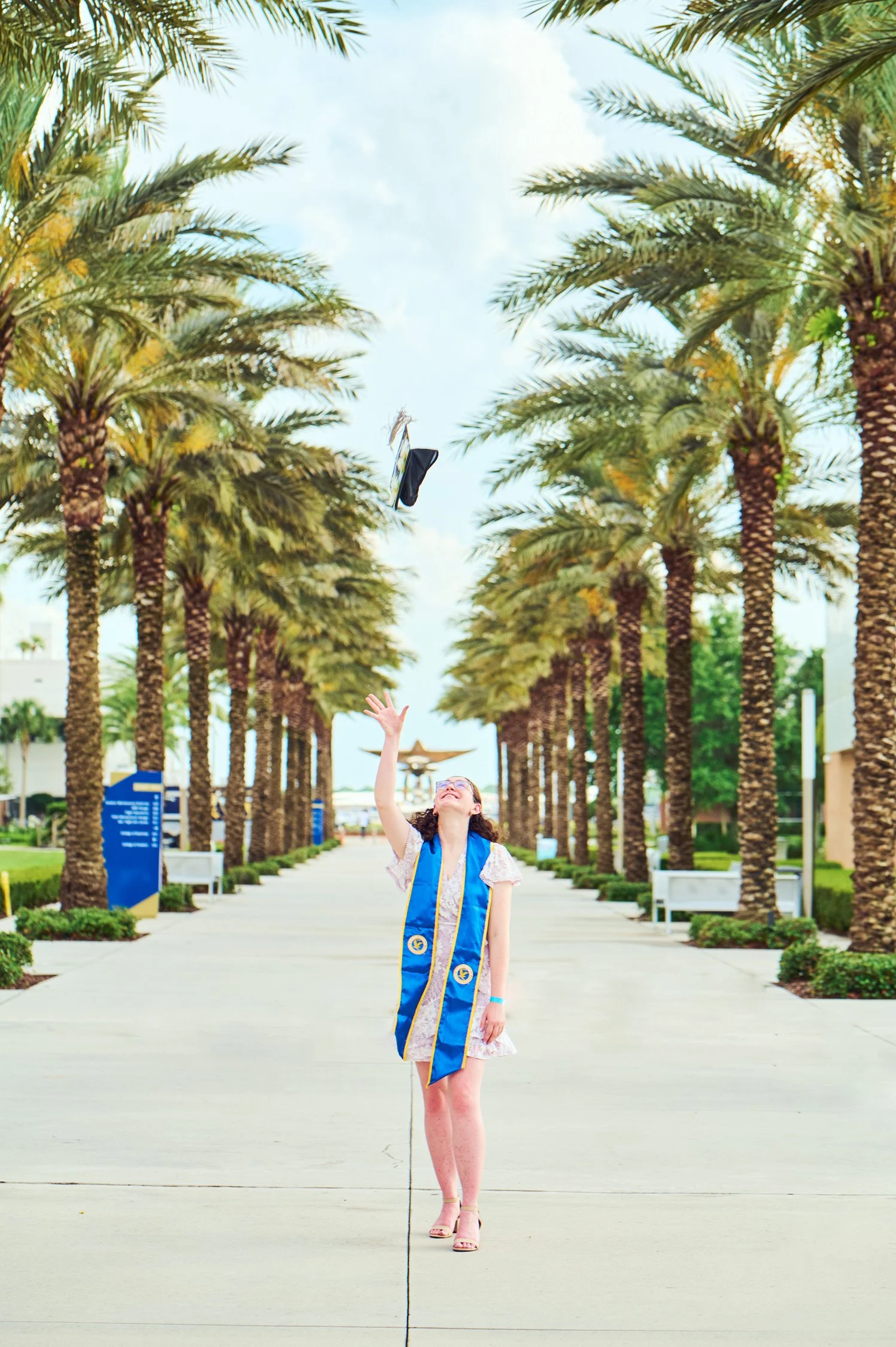 A young woman in a graduation cap and gown dress throws her cap into the air on a wide, palm-lined walkway with benches and trees.
