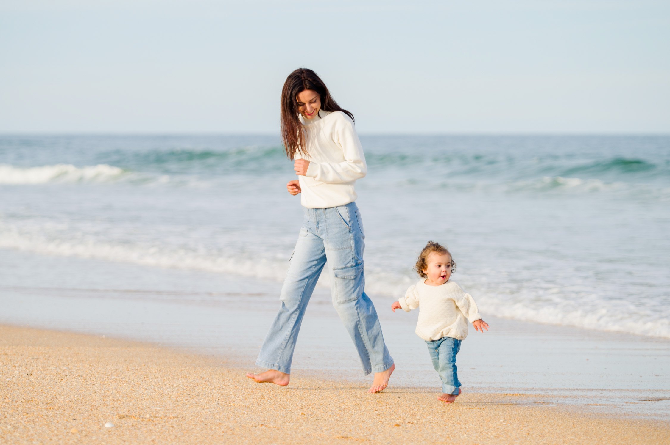 A woman and a young child walking on the sandy beach near the ocean, smiling and enjoying the moment.