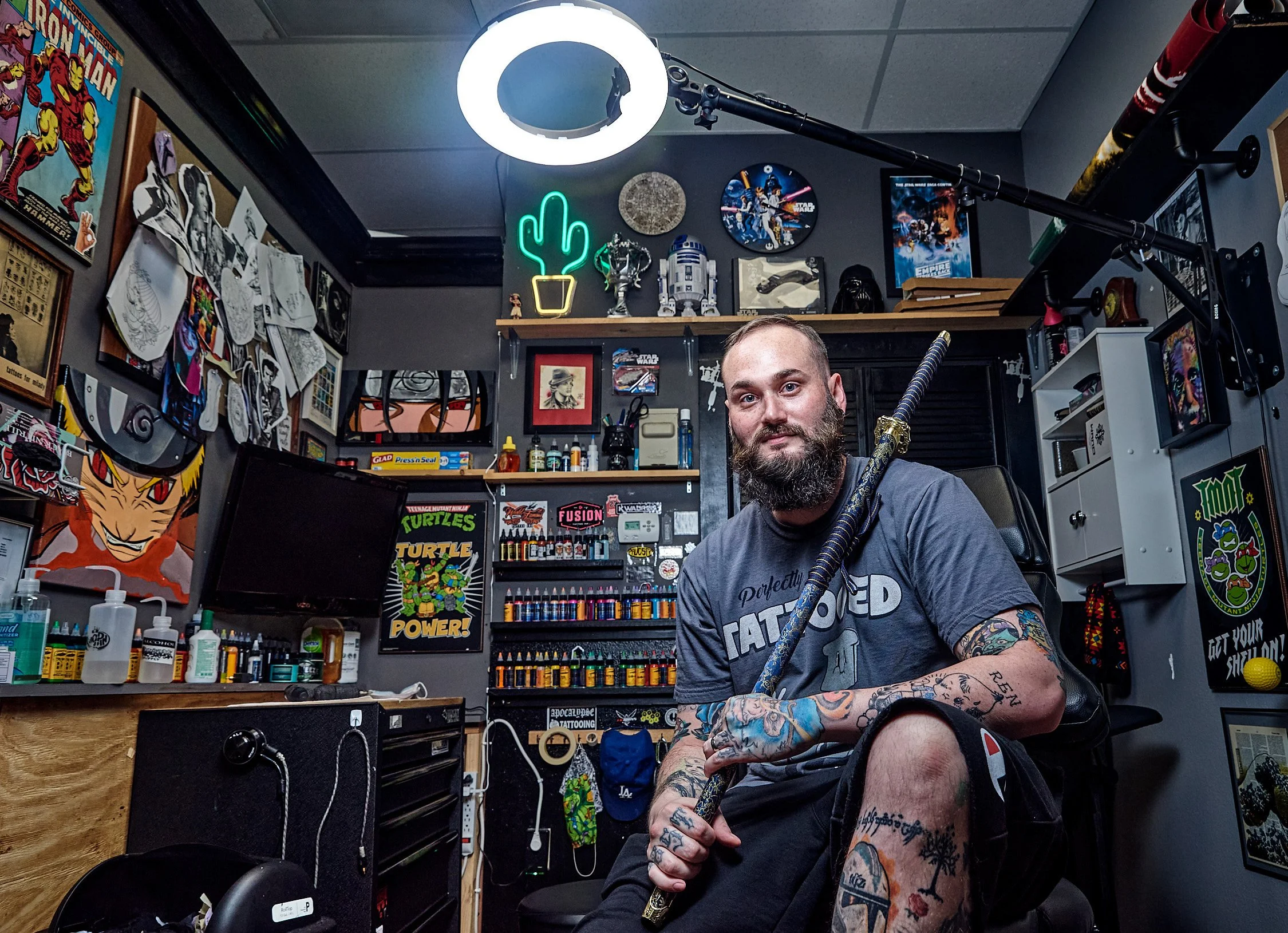 A man with tattoos on his arms and beard sitting in a tattoo studio, holding a katana sword, surrounded by tattoo art and decorations on the walls, including posters of turtles, comic characters, and neon lights.