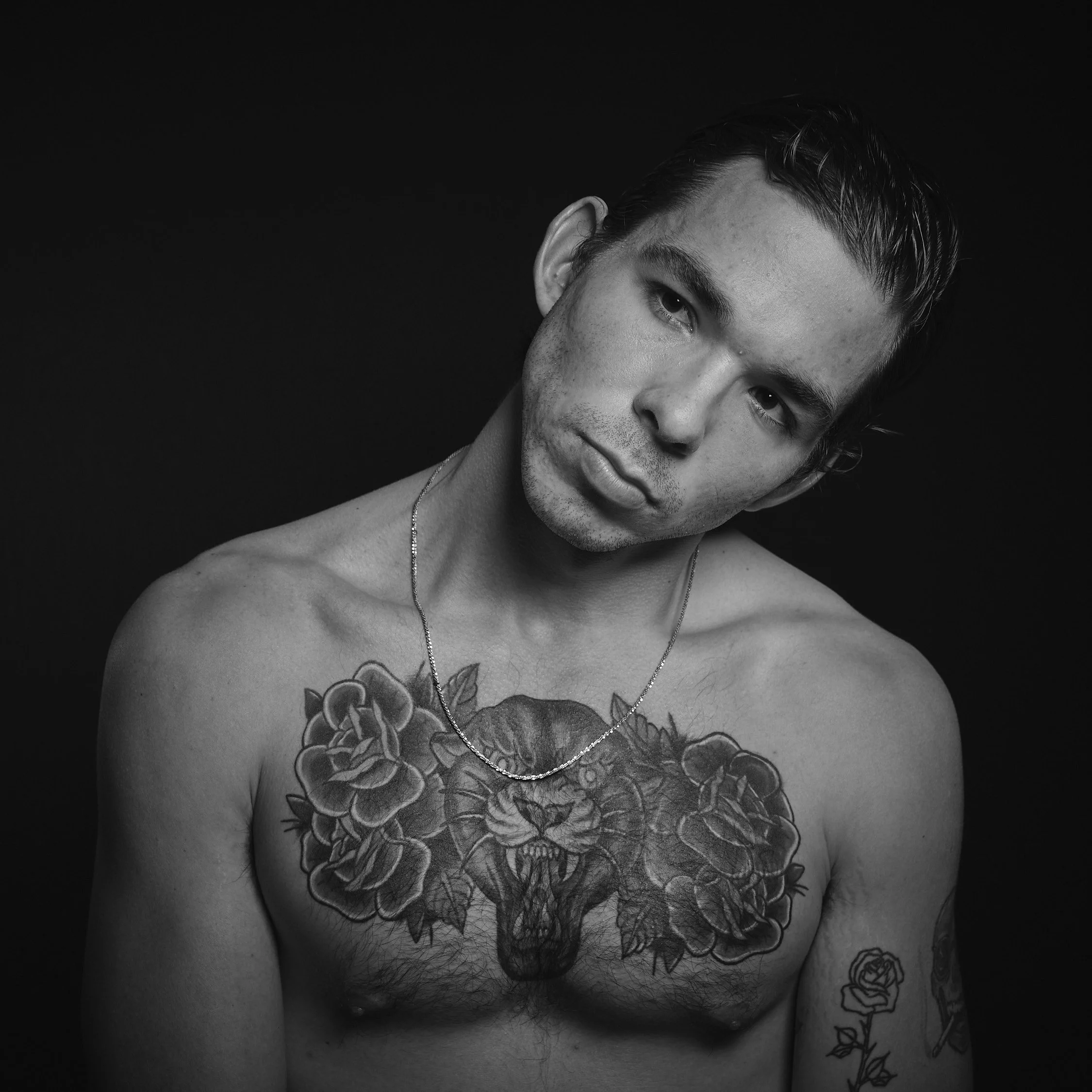 A shirtless man with tattoos, including a tiger head on his chest, a rose on his upper arm, and another rose on his forearm, looking at the camera with a neutral expression against a dark background.