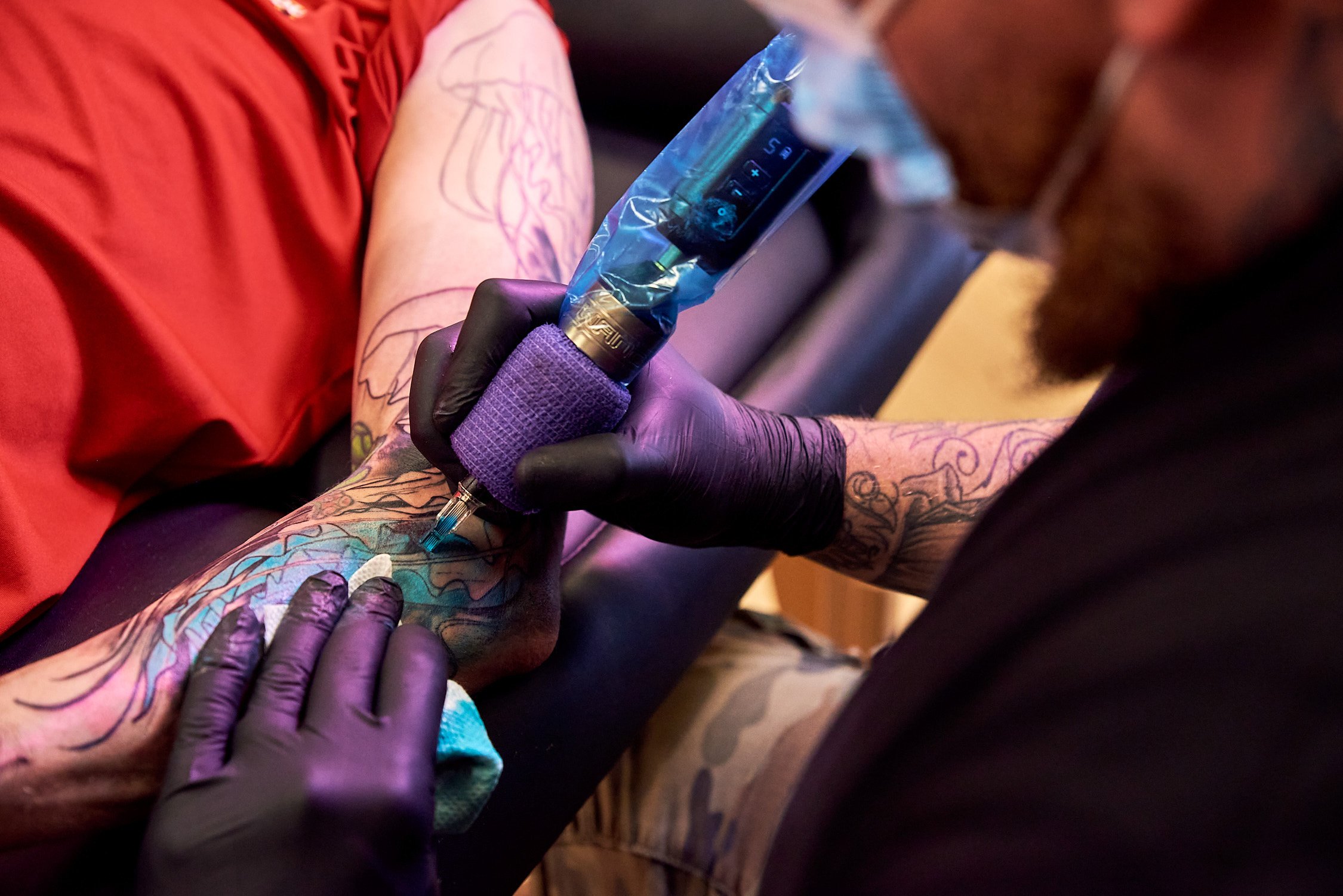 A tattoo artist with gloves tattooing a colorful design on a person's arm using a tattoo machine, with the person wearing a red shirt.