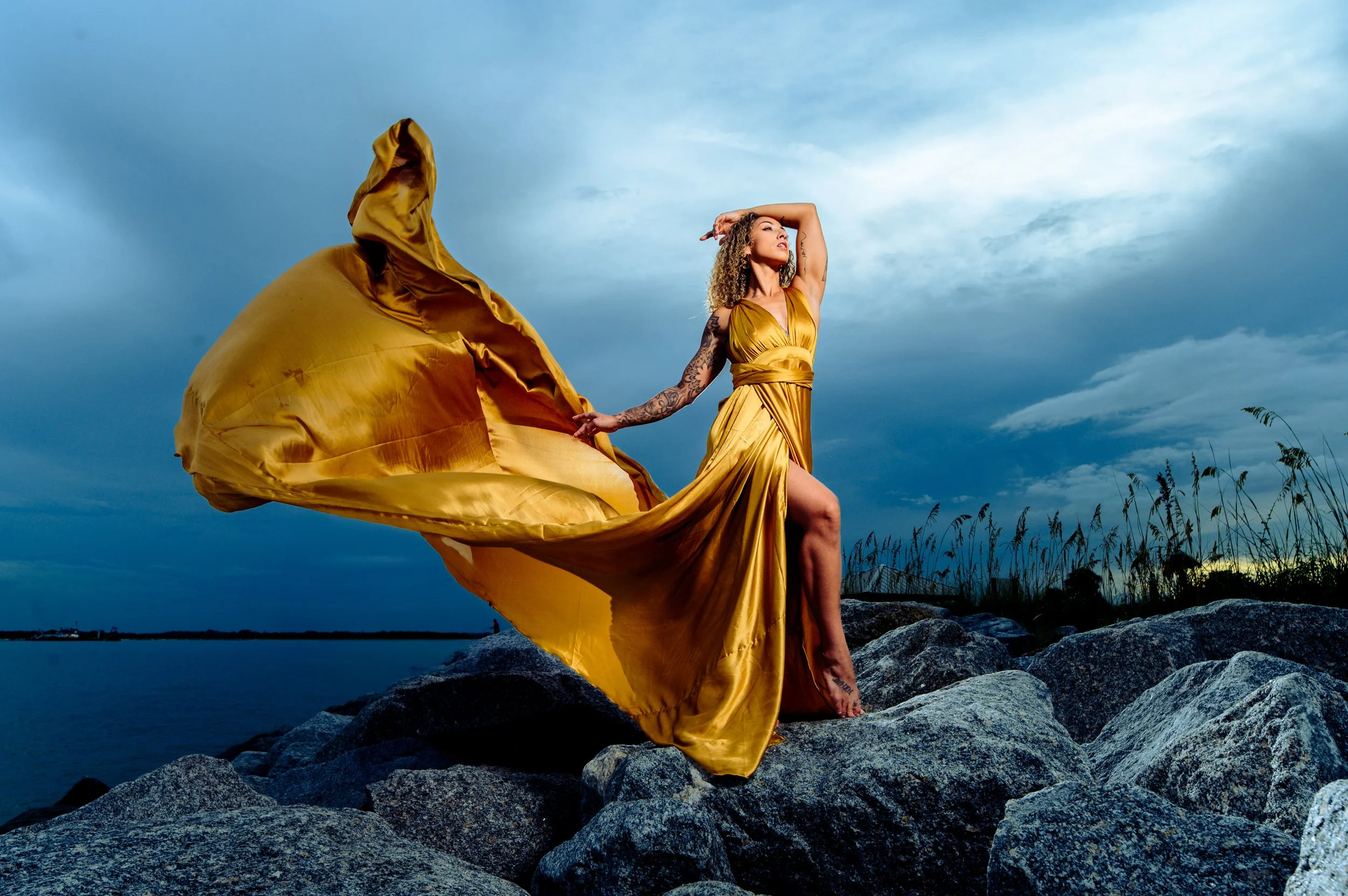 Woman wearing a gold dress standing on rocks near water with a cloudy evening sky in the background.