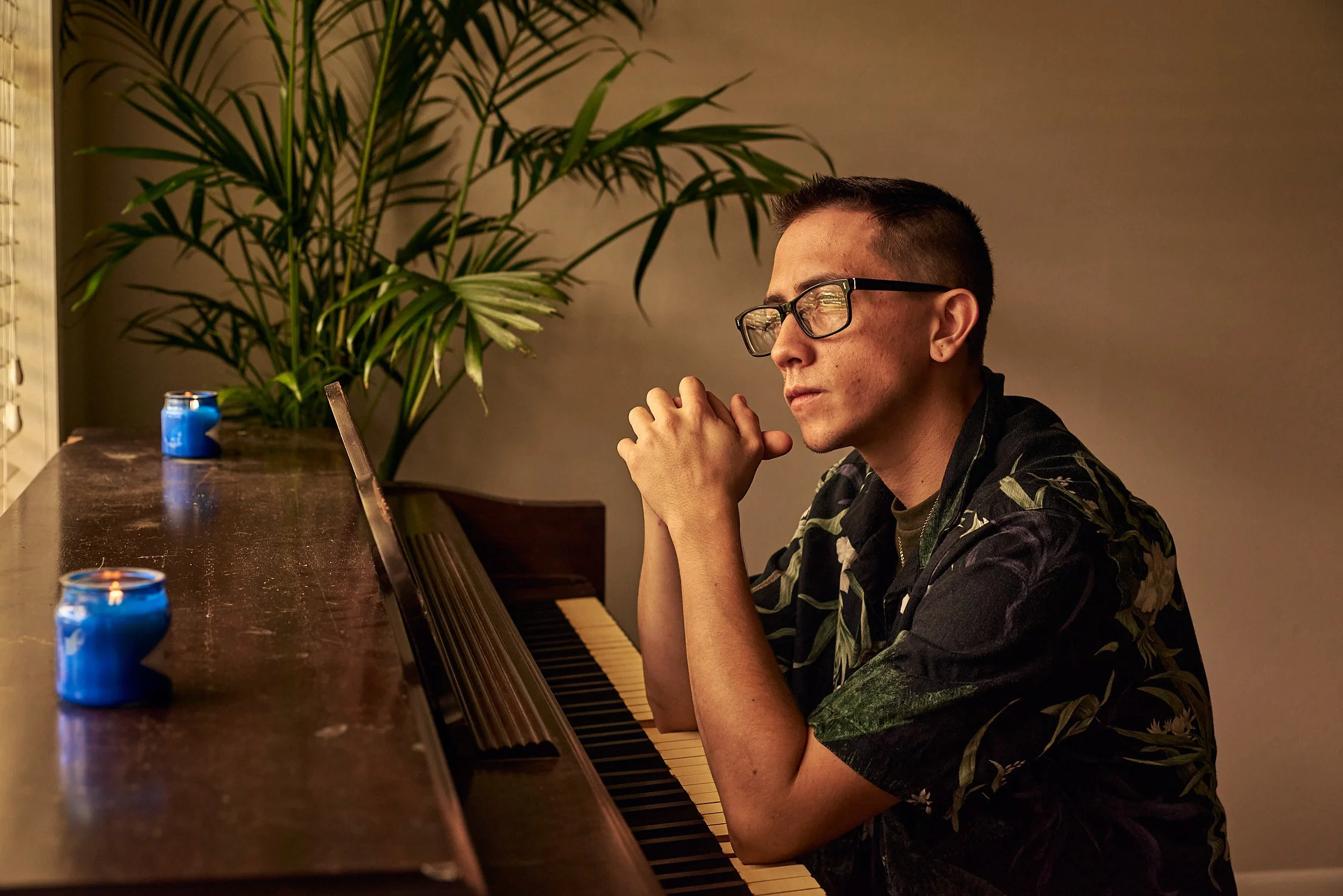 A man with glasses and a short haircut sitting at a piano, with his hands clasped and eyes closed, in a room with green plants and two blue candles on the piano.