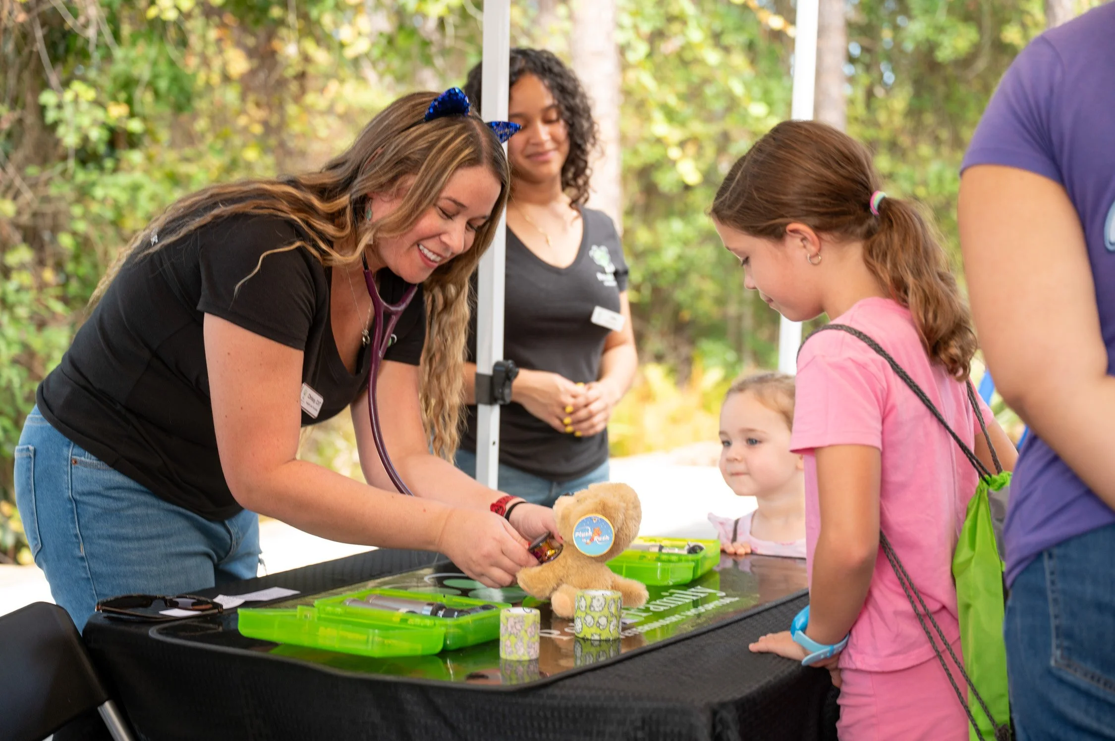 A woman with a stethoscope around her neck is showing a stuffed bear to two young girls at an outdoor event, with another woman in the background. The scene is set in nature with trees in the background.