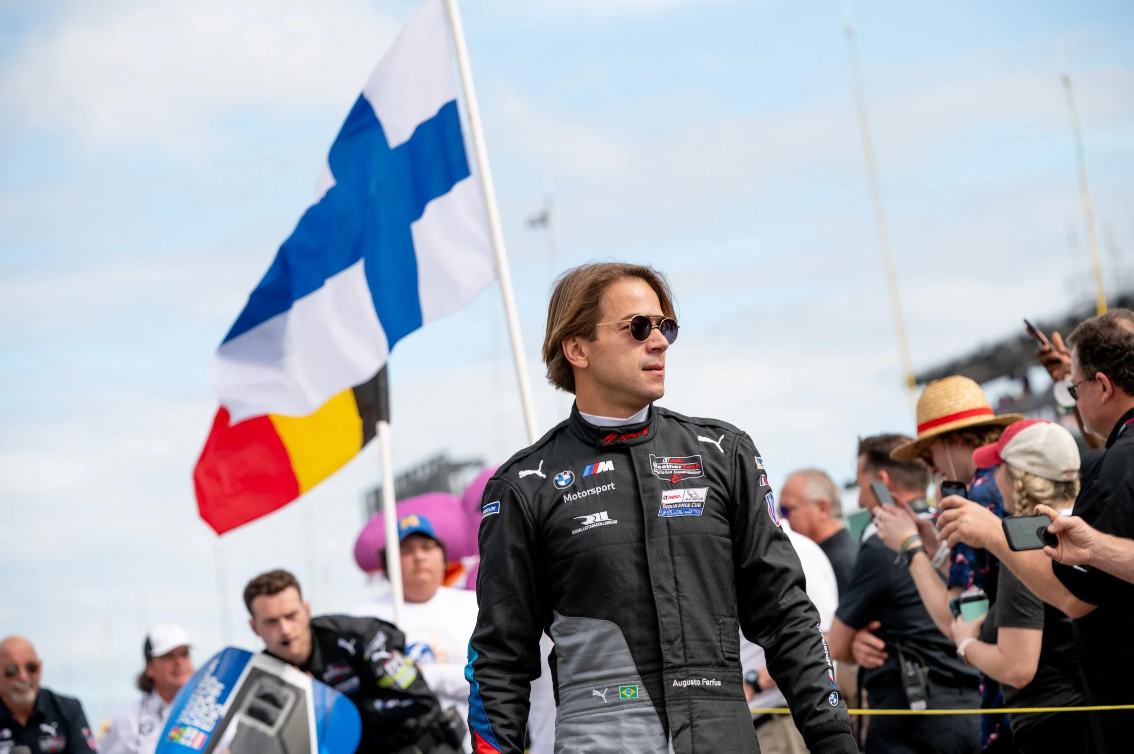 A man in a black racing suit with sponsors' logos, sunglasses, and medium-length hair, stands at a racing event holding a Finnish flag, with a crowd and some people taking pictures in the background.