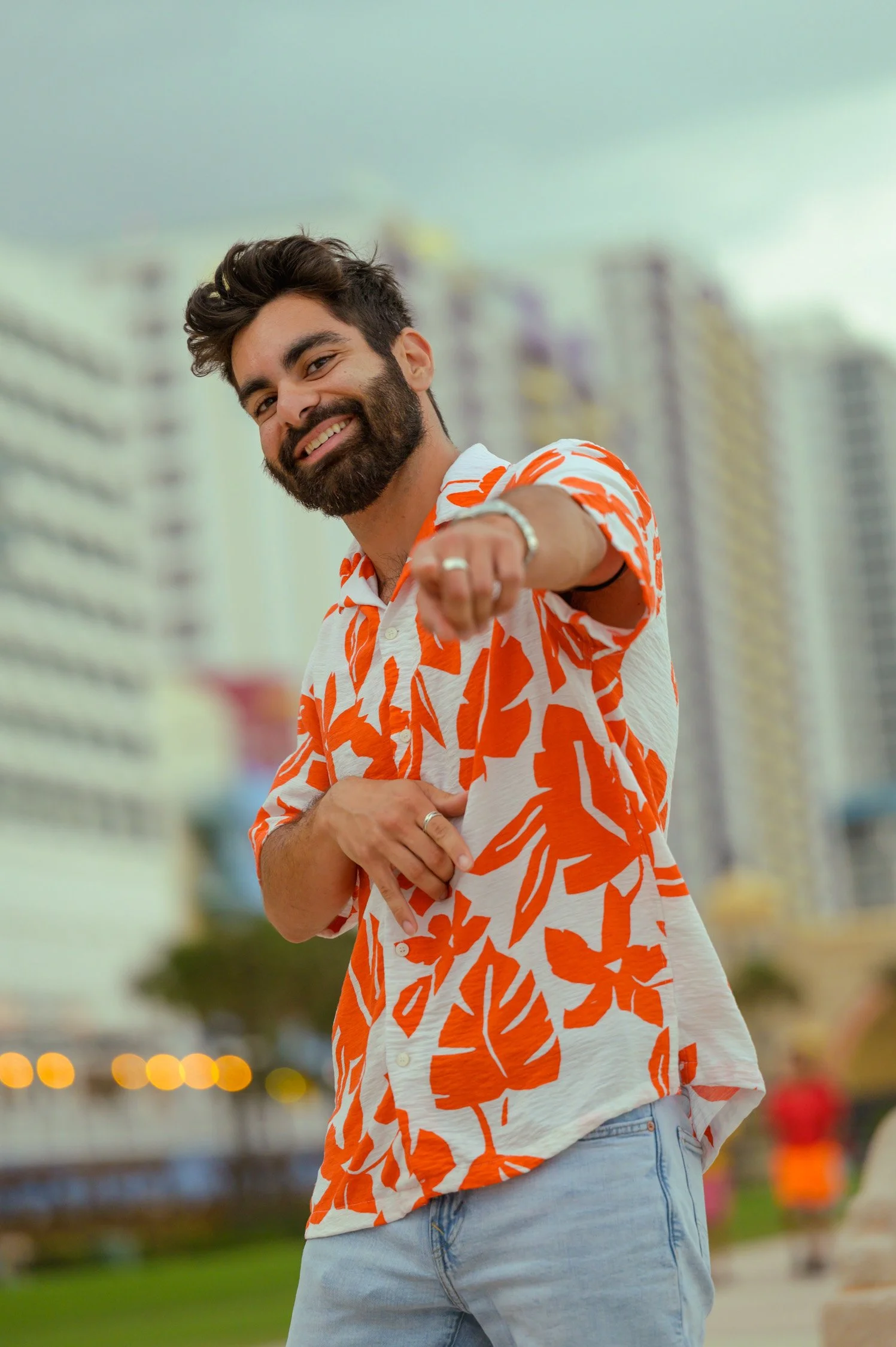 A smiling man with a beard wearing a brightly patterned orange and white shirt, pointing towards the camera outdoors in an urban setting.
