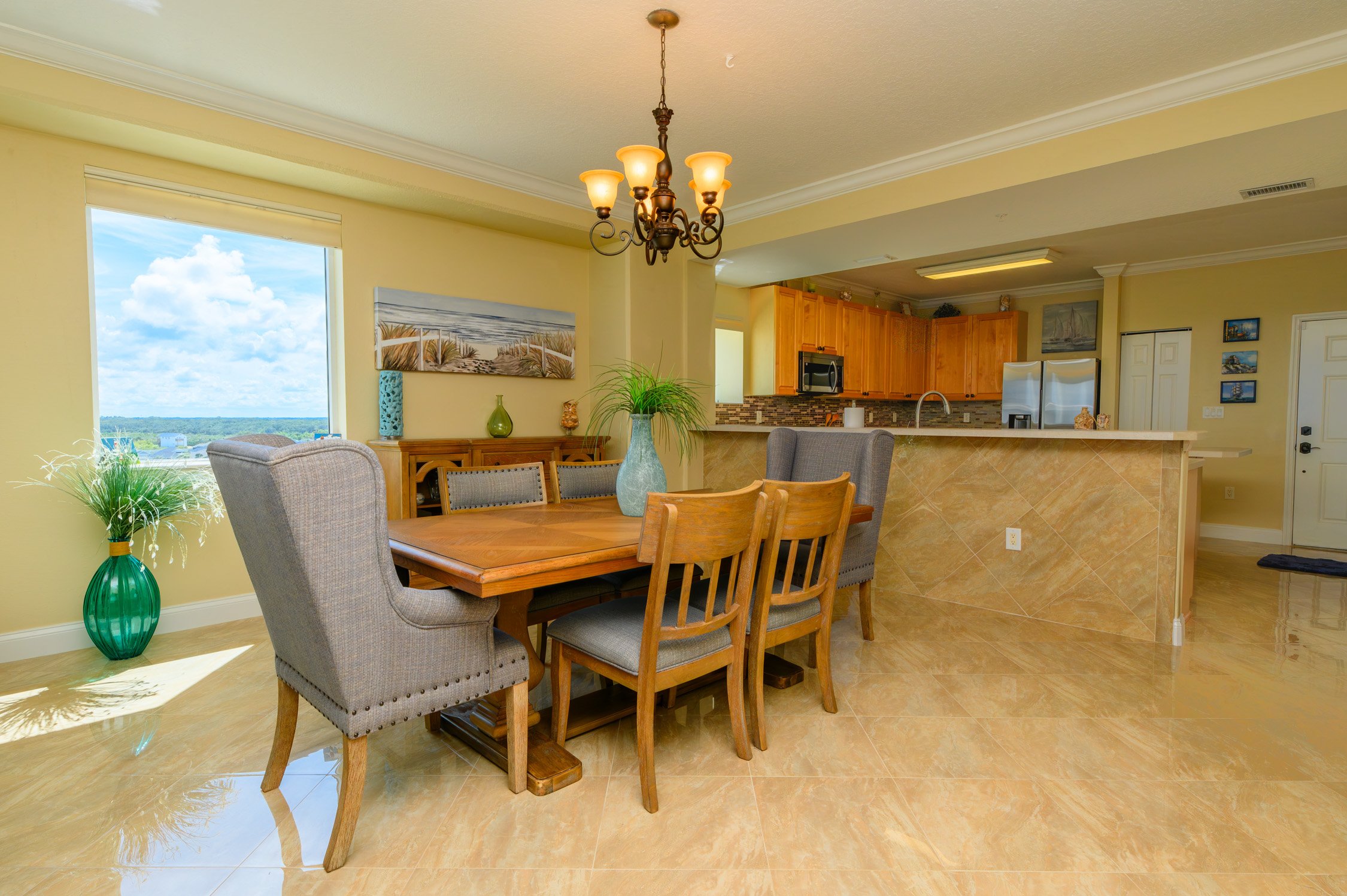 Dining area with a wooden table, mixed gray and wooden chairs, large window with a view, and colorful plants, adjacent to a kitchen with wooden cabinets and a tiled counter.