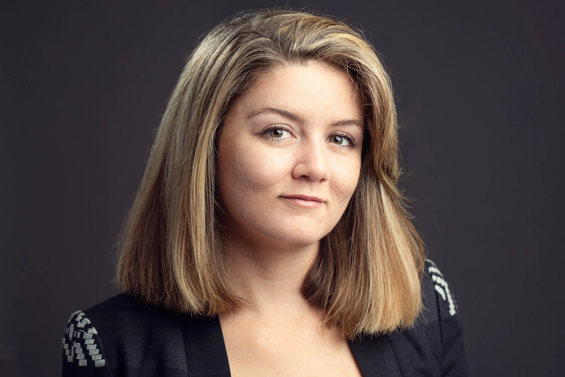 A woman with shoulder-length light brown hair and blue eyes, wearing a black top with white details on the shoulders, against a dark gray background.