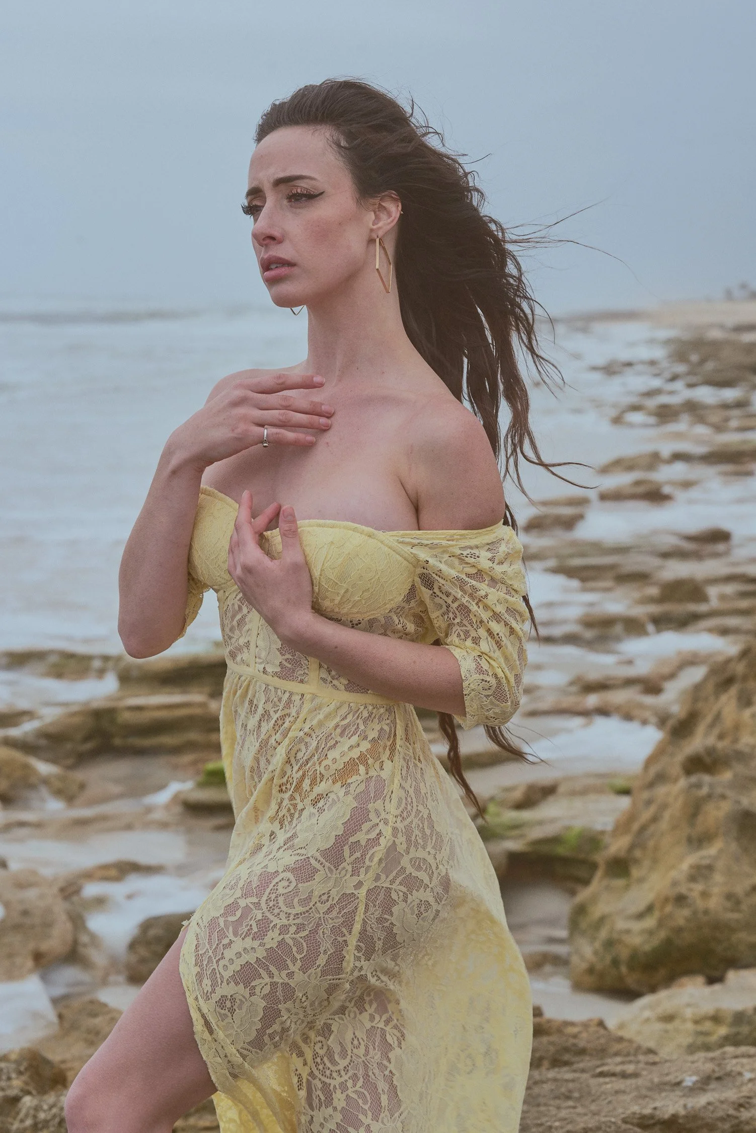 A woman in a yellow lace dress standing on a rocky beach, looking thoughtfully into the distance with her hair blowing in the wind.