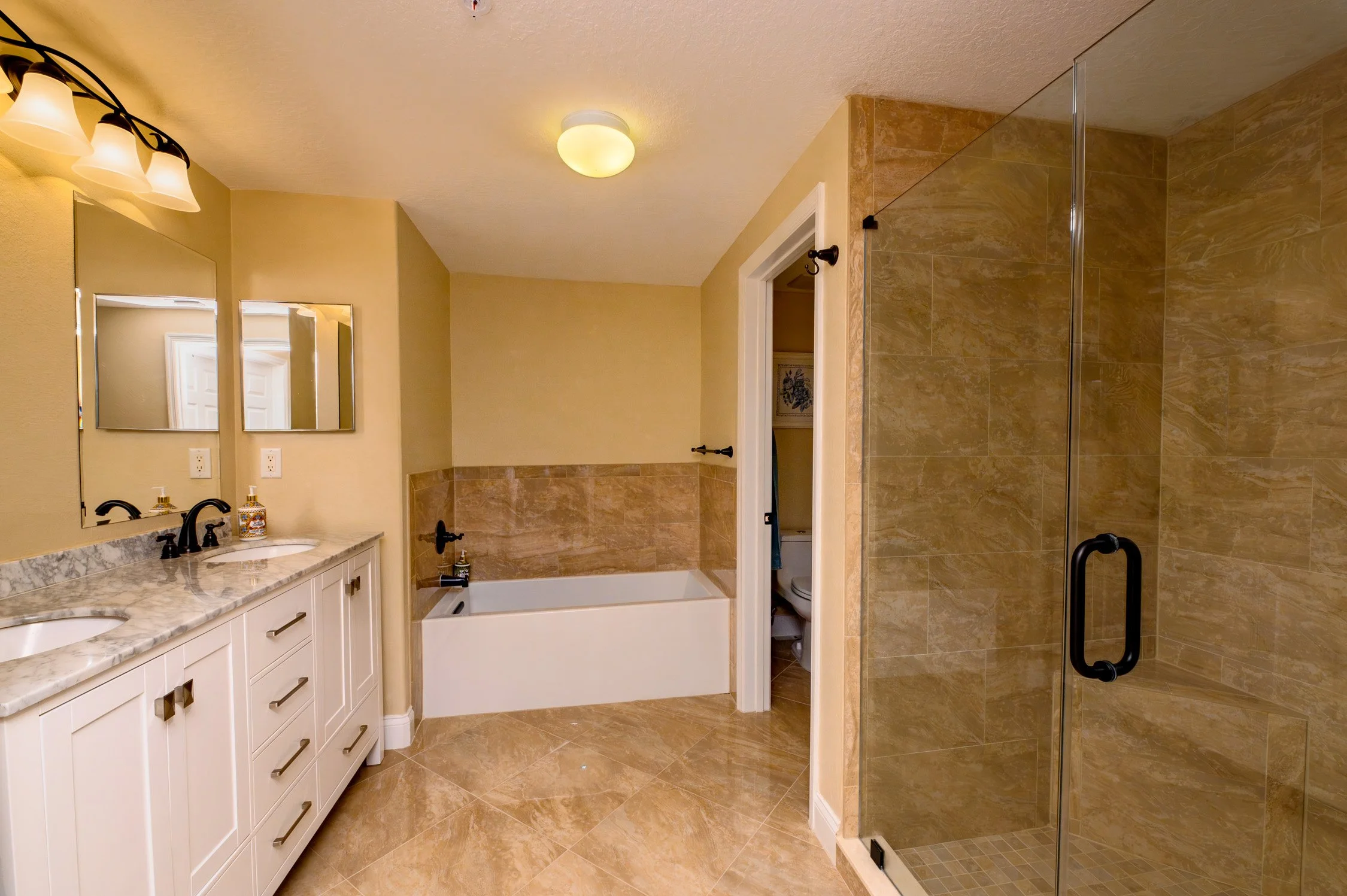 A bathroom with dual sinks and mirrors on a marble countertop, a bathtub with a partial wall tile surround, a toilet in a small room, and a glass-enclosed shower with tan stone tiles.