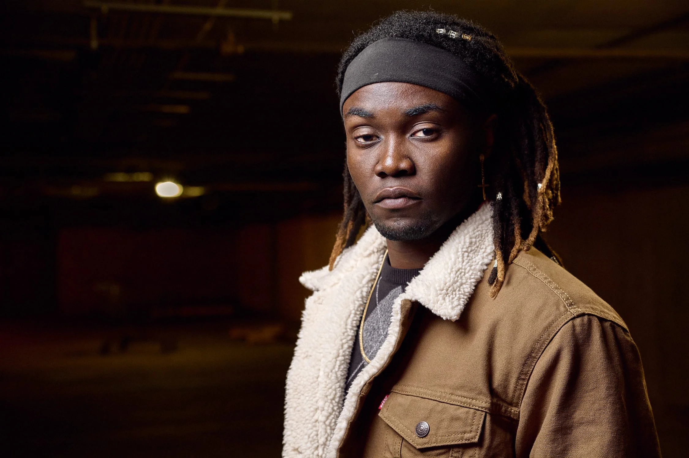 A young man with dreadlocks wearing a black headband, a tan jacket with a shearling collar, and gold cross earring, standing in a dimly lit environment with serious expression.