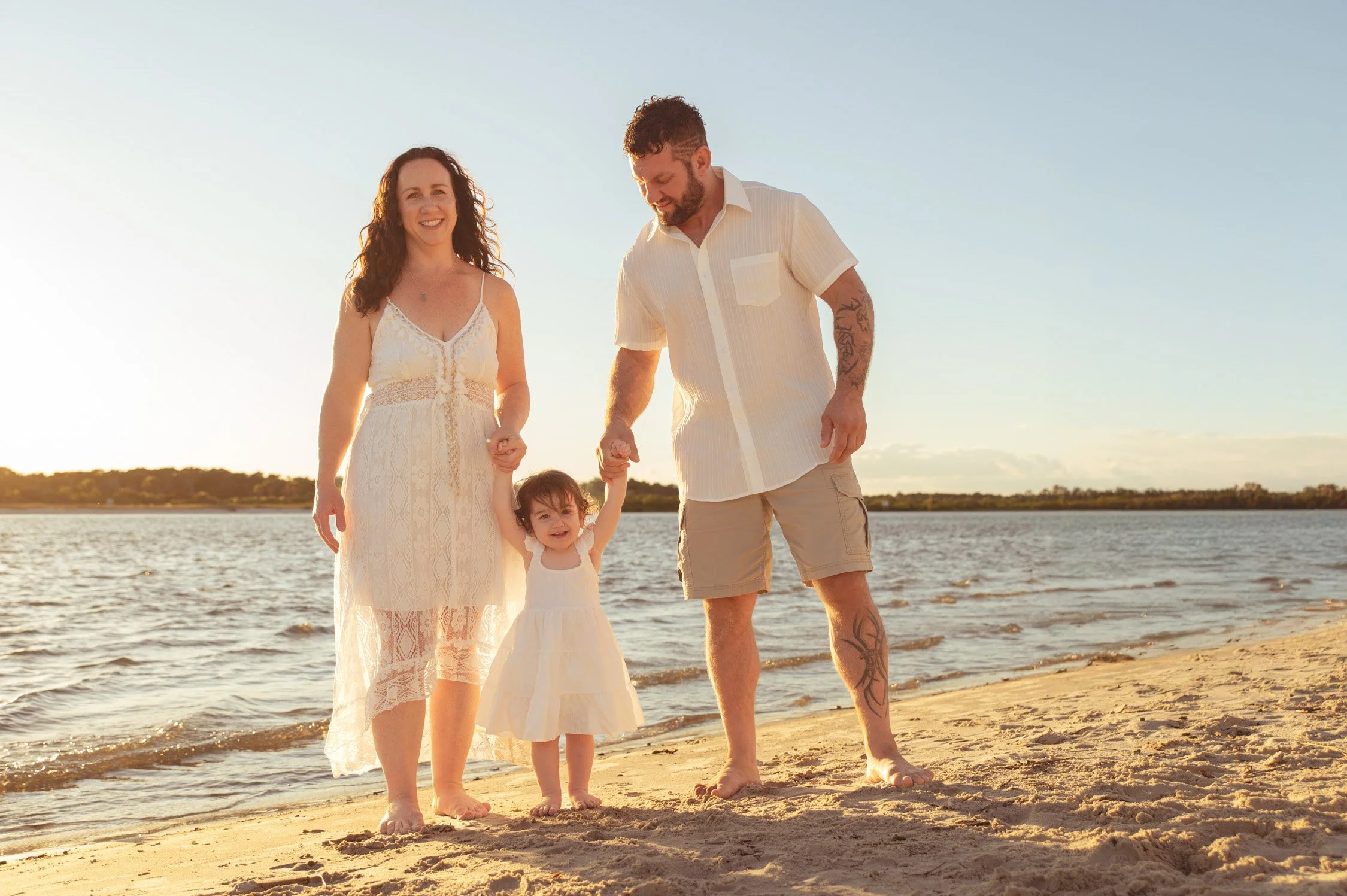 A family of three, a woman, a man, and a young girl, walking barefoot on the beach at sunset. The woman is wearing a white lace dress, the man is in a white short-sleeve shirt and beige shorts, and the girl is in a white dress, smiling and holding ha