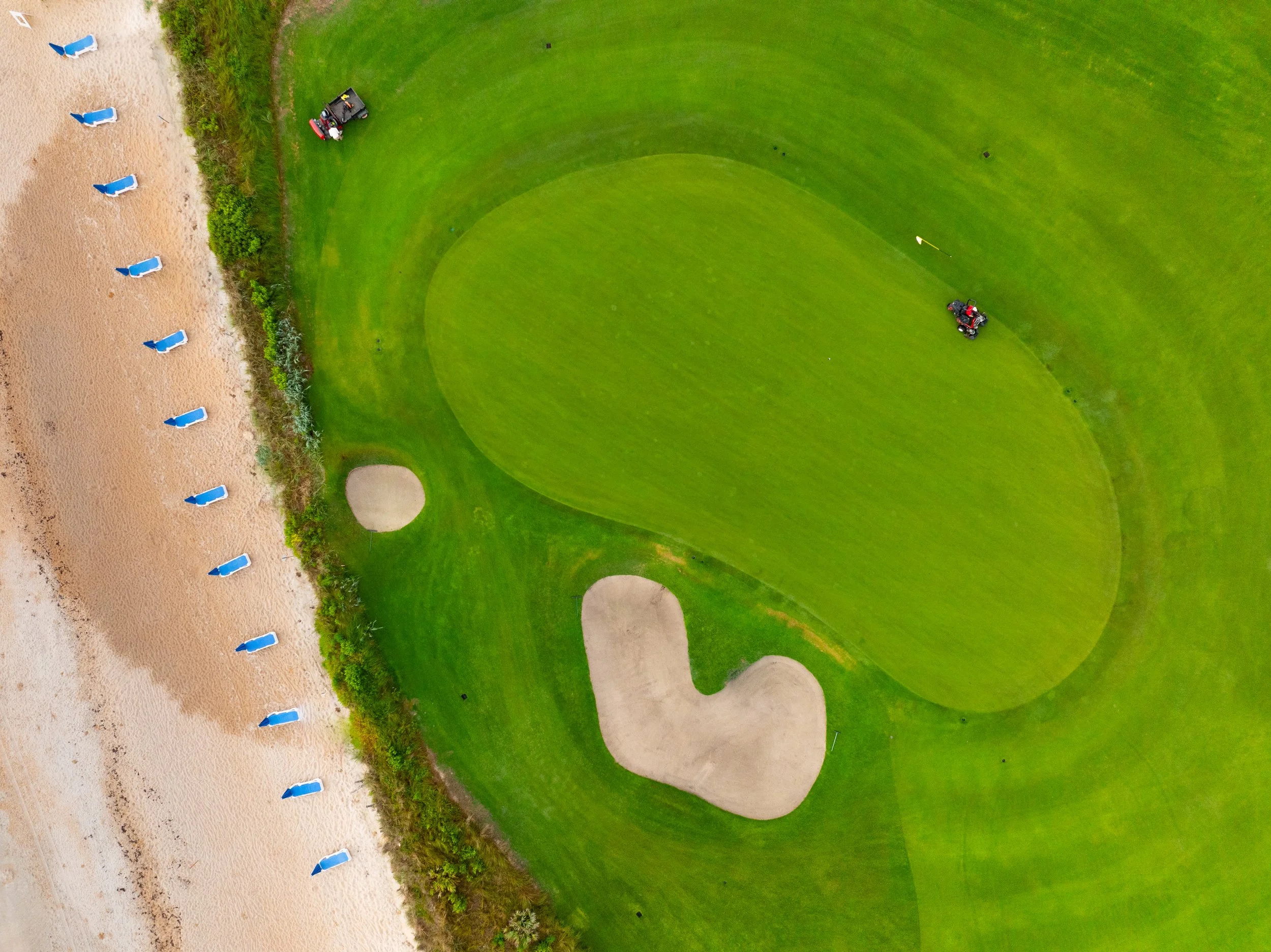 Aerial view of a golf course with sand bunkers and a putting green next to a sandy beach with blue lounge chairs