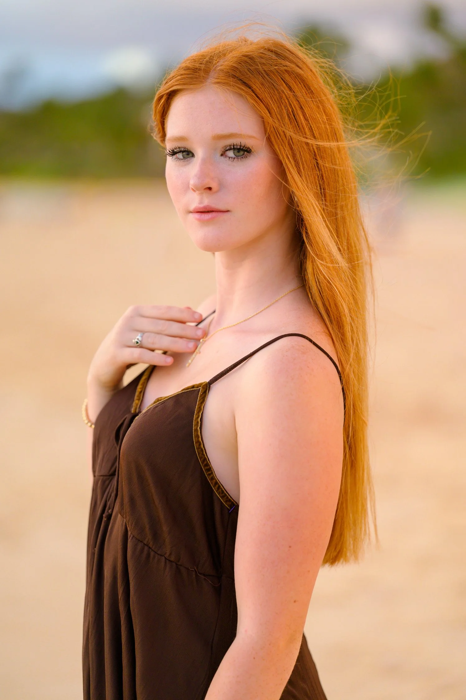 A young woman with long red hair, wearing a brown spaghetti strap dress, standing outdoors with a blurred background of green trees and sky.