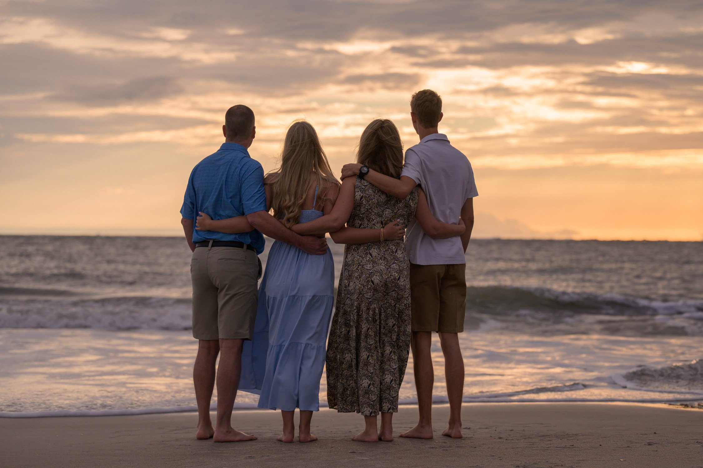 Four people standing arm in arm on a beach at sunset, facing the ocean.