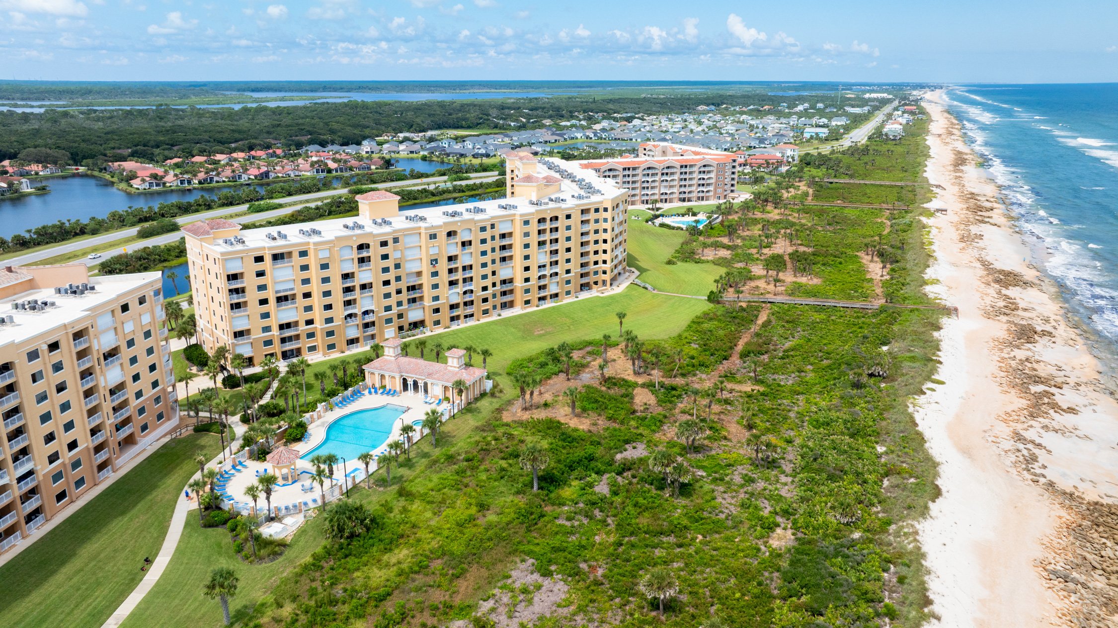 An aerial view of a beachfront resort with multiple beige buildings, a swimming pool, green lawns, trees, and a sandy beach along the ocean.