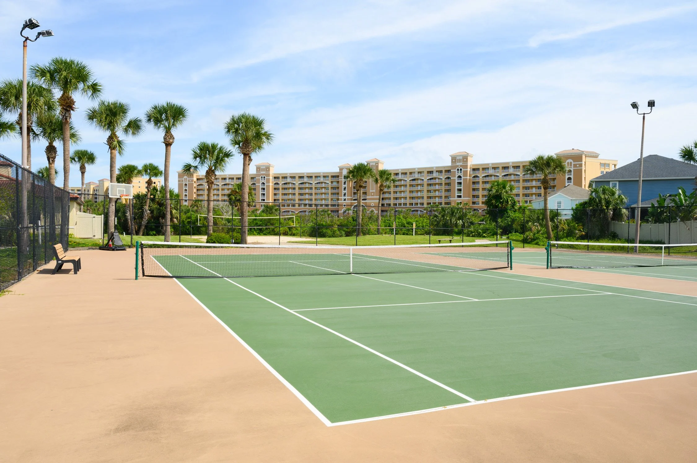 Empty tennis court with green surface, white lines, and a net, surrounded by palm trees, apartment building, and blue sky with clouds.