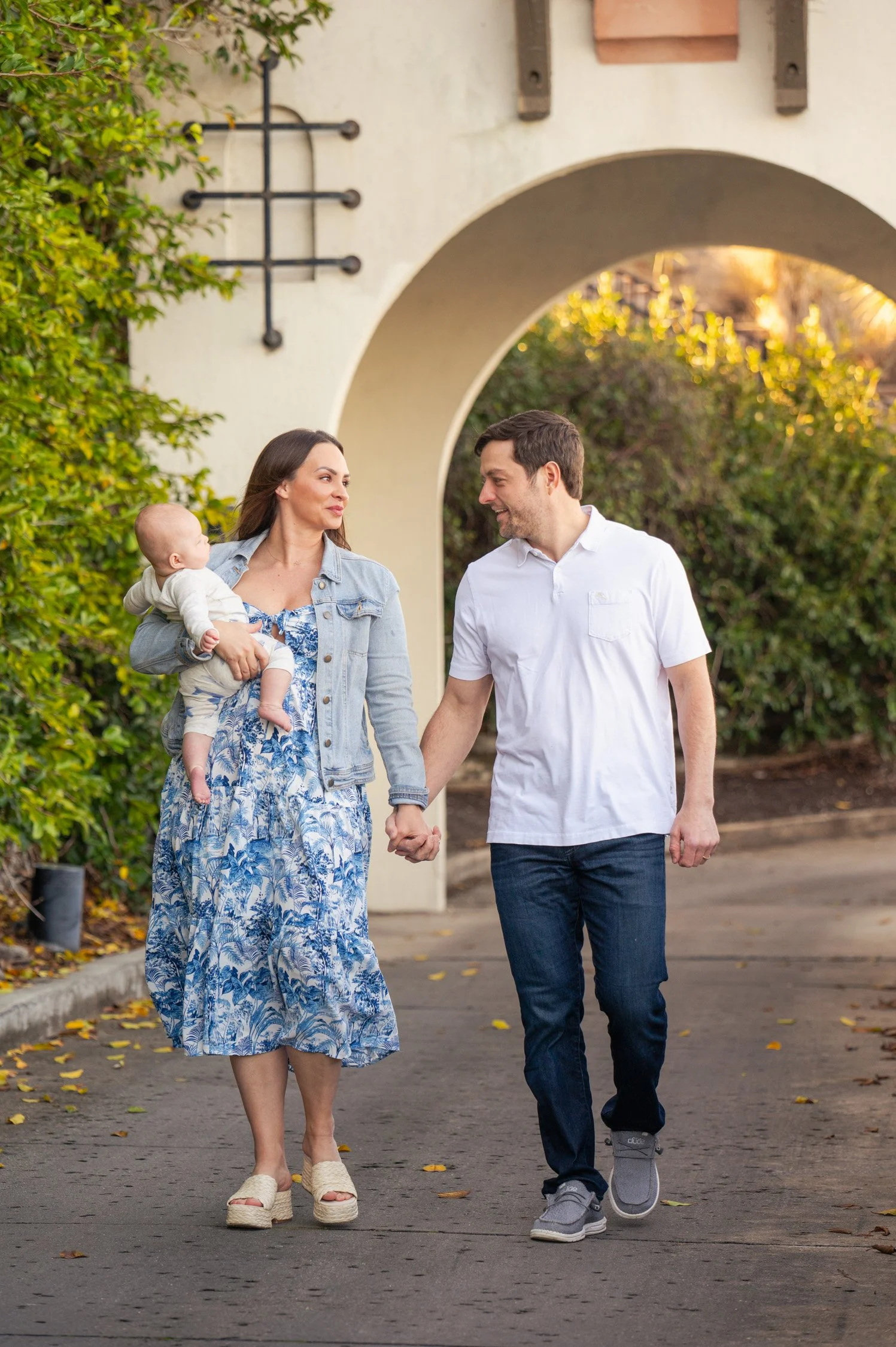 A family of three, a woman holding a baby and a man, walk hand in hand outdoors near an archway with greenery and trees in the background.