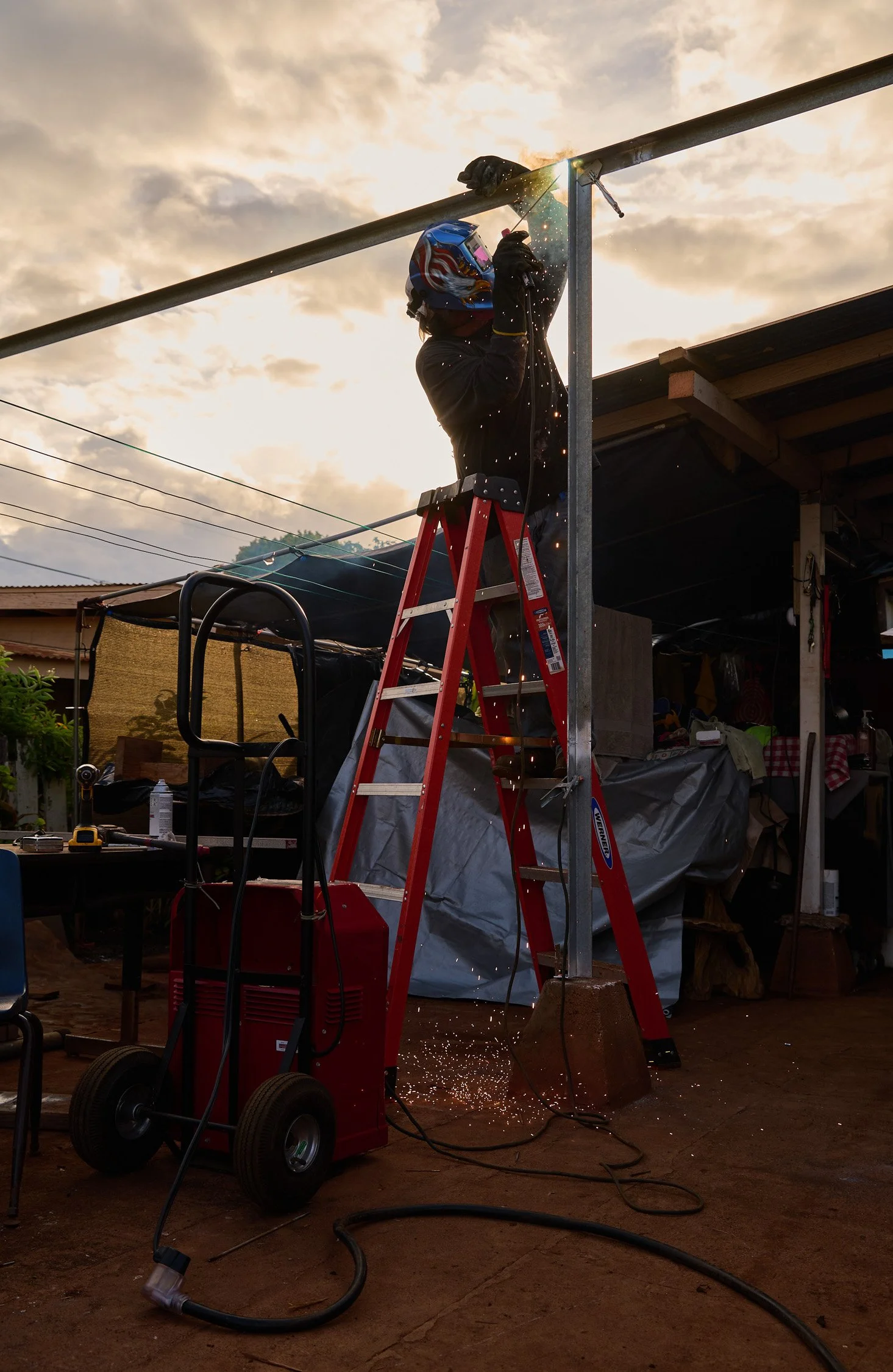 Person welding on a metal structure outdoors at sunset, with sparks flying, on a red ladder.