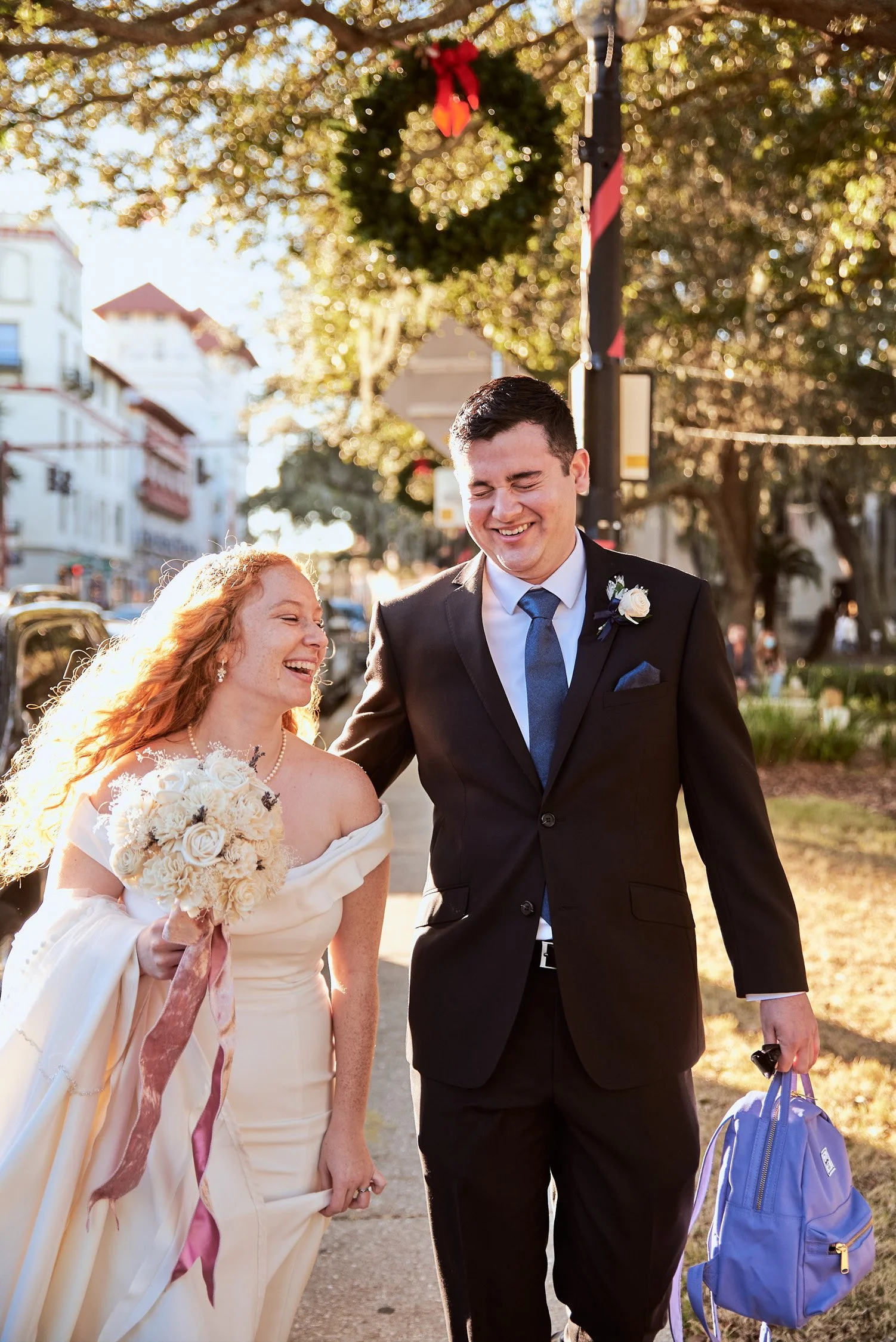 A smiling bride and groom walk outside during sunset, with the bride holding a bouquet, and the groom carrying a blue bag, surrounded by trees and buildings.