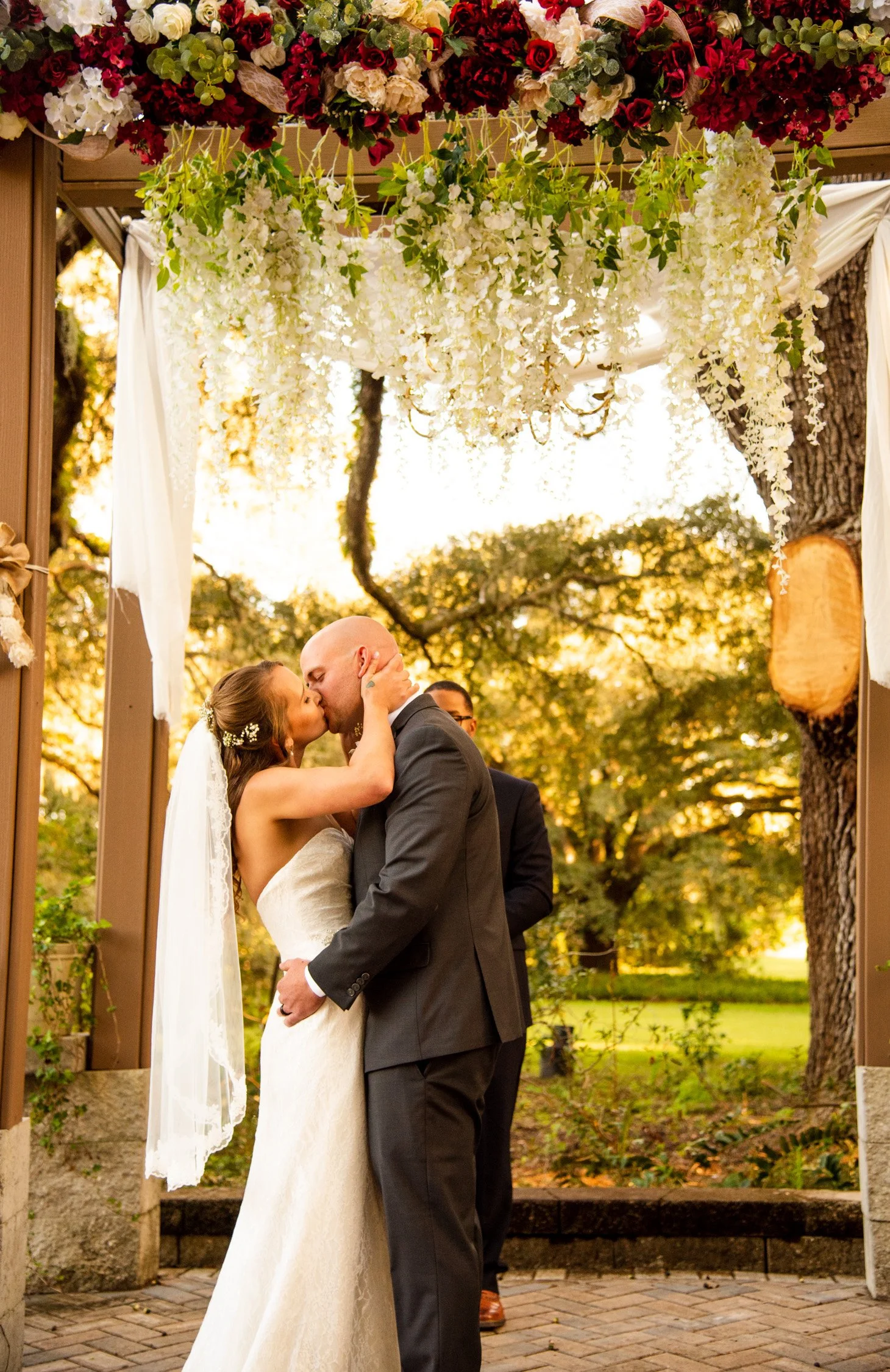 A bride and groom share a kiss during their outdoor wedding ceremony under a floral arch with white and red flowers, greenery, and hanging white and red blooms, set against a backdrop of trees and warm sunlight.