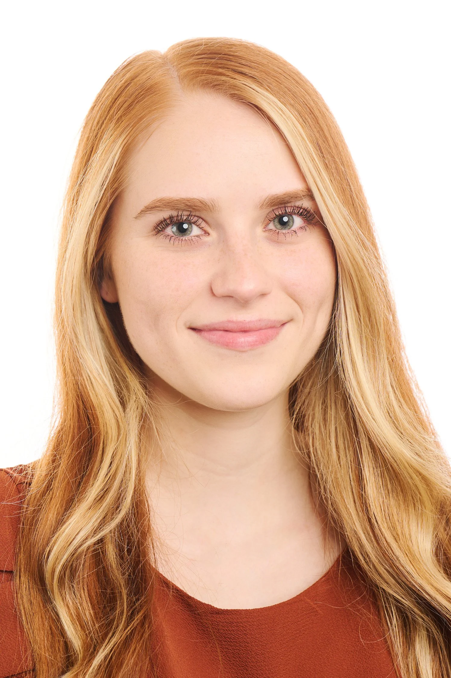 Close-up portrait of a young woman with red hair, blue eyes, and a gentle smile, wearing a rust-colored top, against a white background.