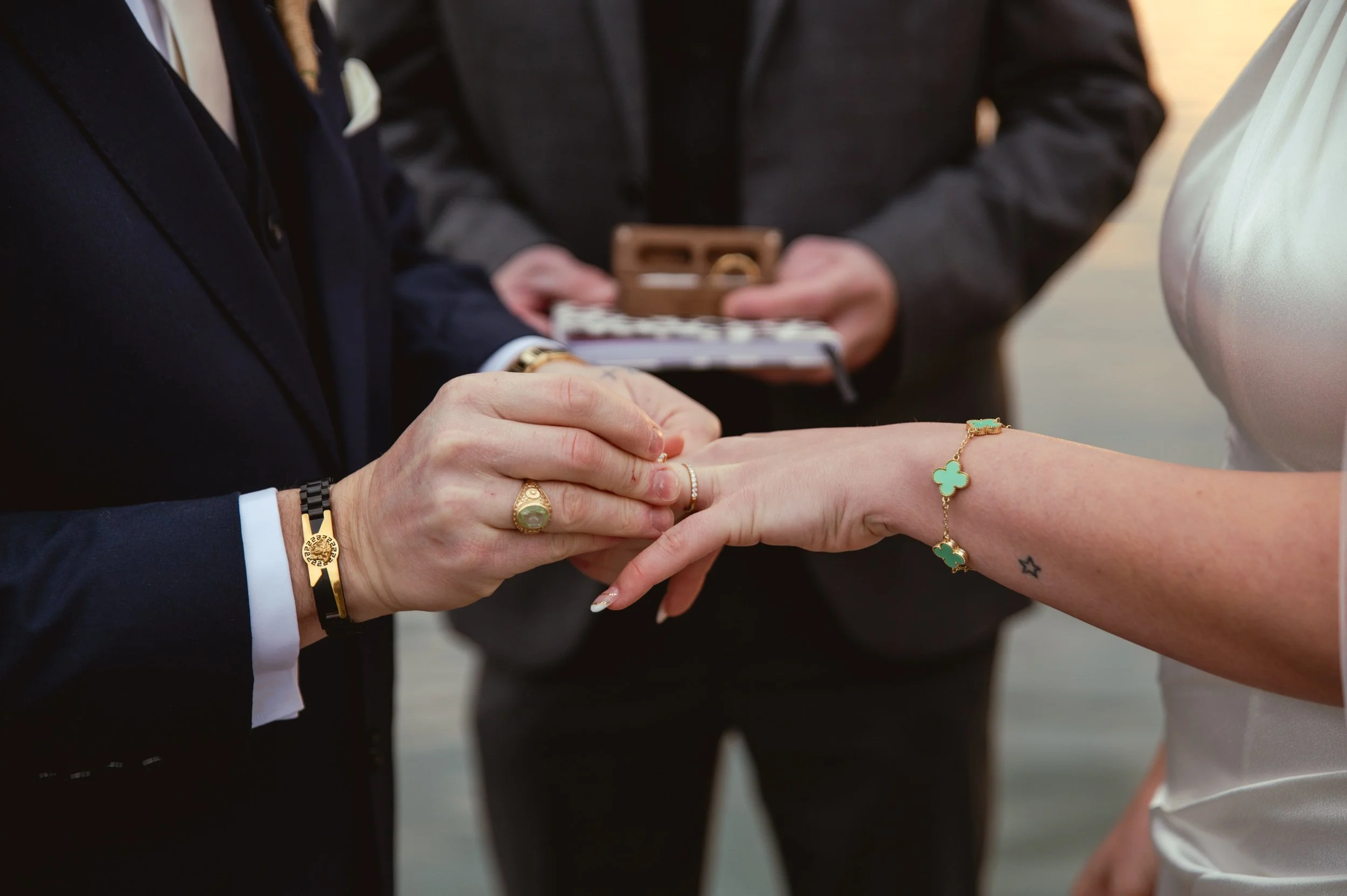 Close-up of a couple exchanging rings during a wedding ceremony by the water, with officiant in background holding a small box.