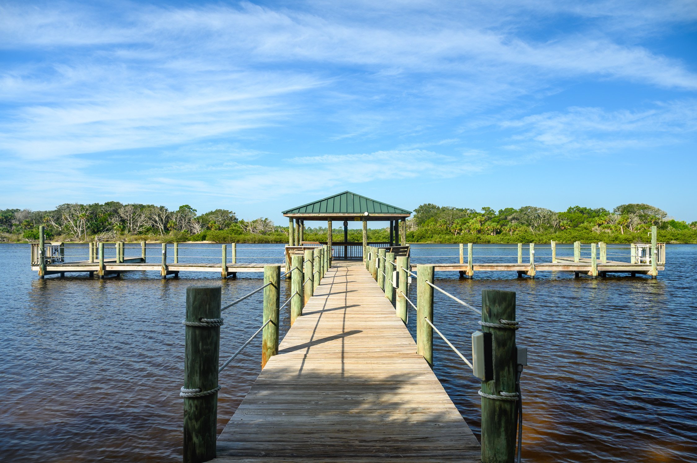 Wooden dock leading to a covered gazebo on the water with trees in the background and a blue cloudy sky.