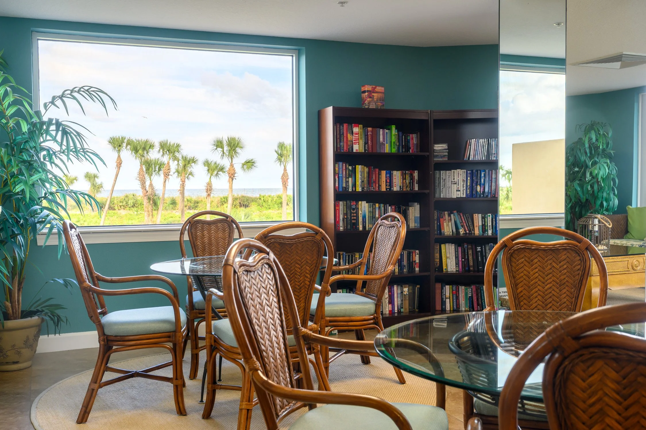 A cozy indoor space with a large window showing palm trees and a beach view, wooden chairs around glass tables, a black bookshelf filled with books, and tropical plants.